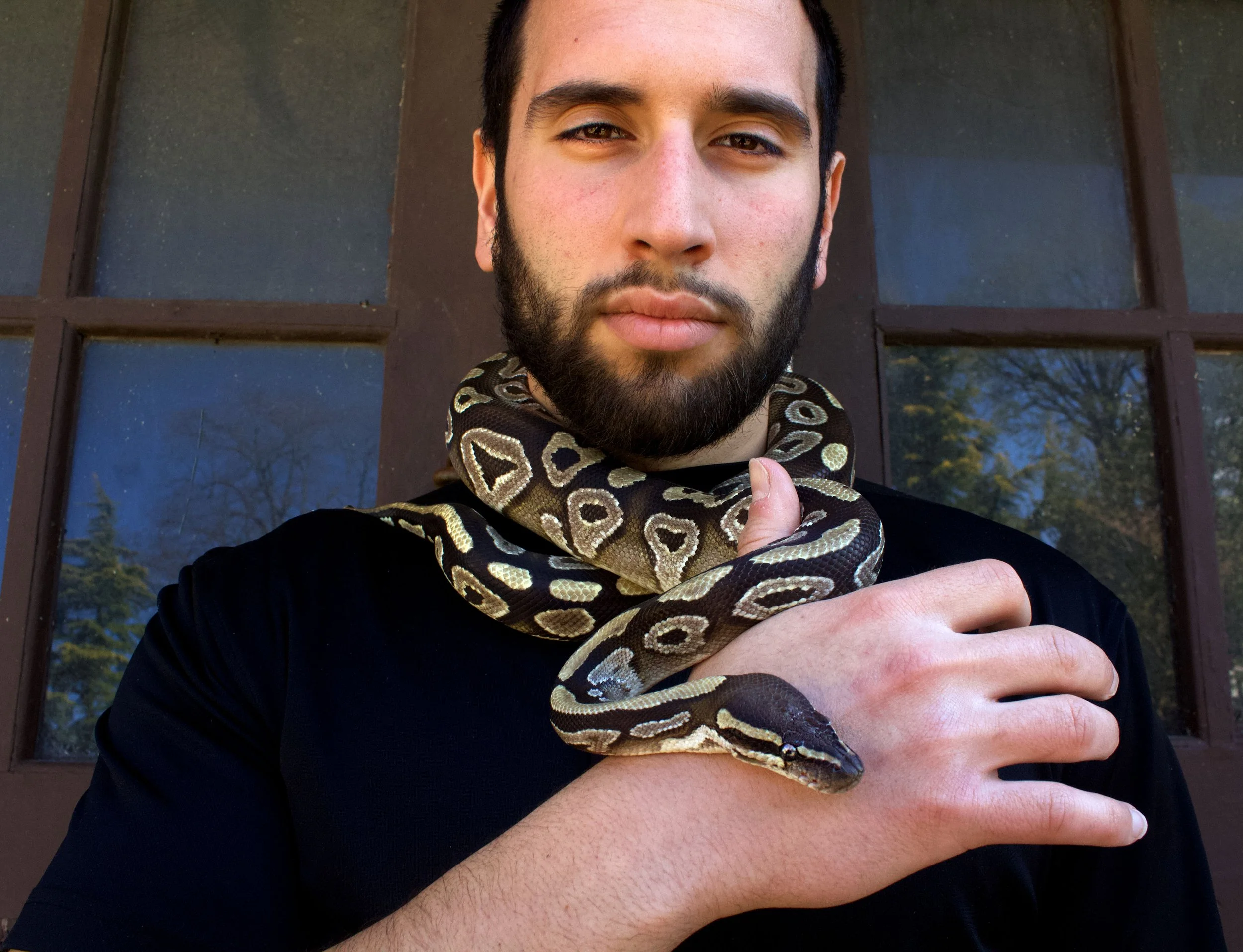 A young man with a beard and mustache taking a selfie outdoors in front of a window. He wears a black shirt and has a small snake around his neck and shoulders, with his right hand visible, showing a relaxed hand gesture.