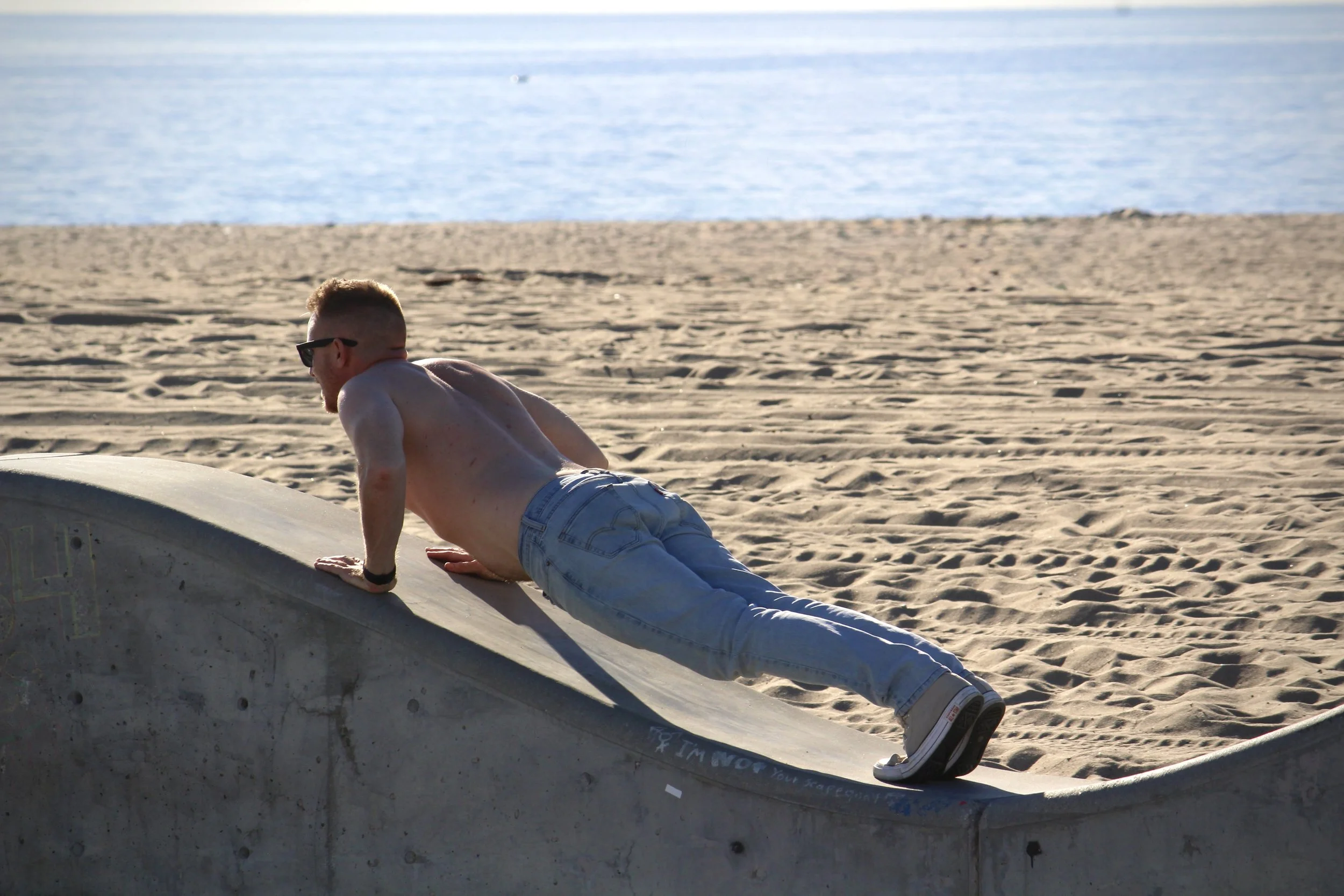 A shirtless man wearing sunglasses and light blue jeans is lying on his side on a sandy beach next to a small white boat, with the ocean in the background.