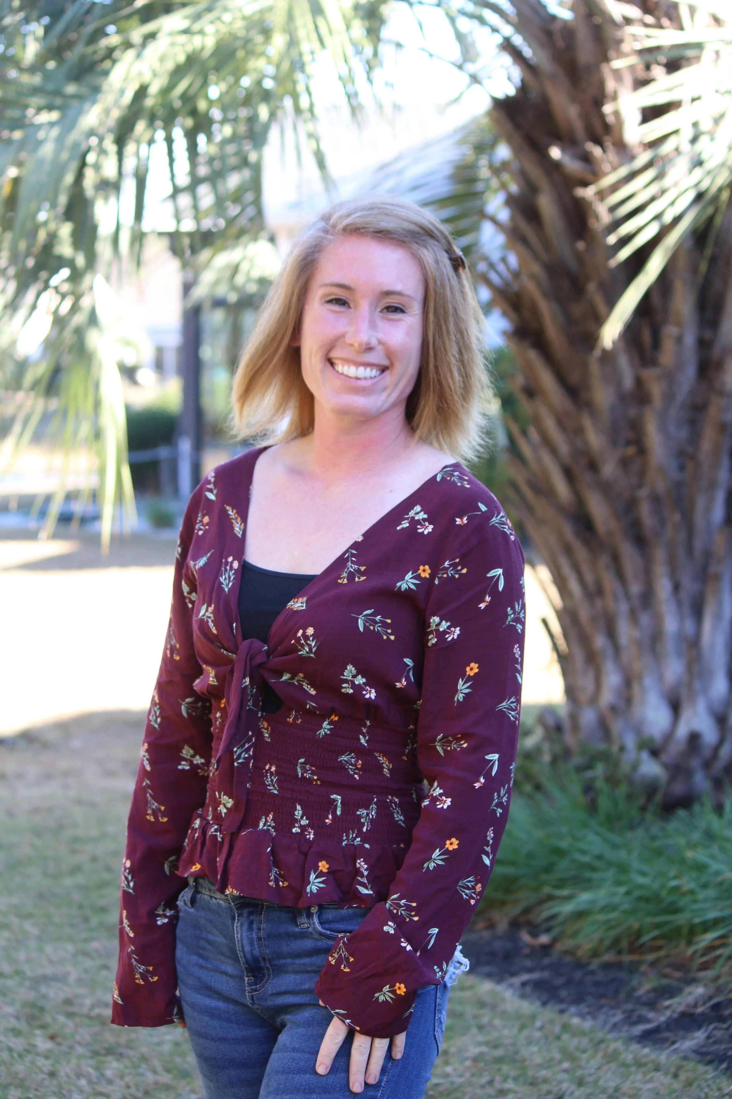 A young woman with blonde hair smiling outdoors near a palm tree, wearing a burgundy floral top and blue jeans