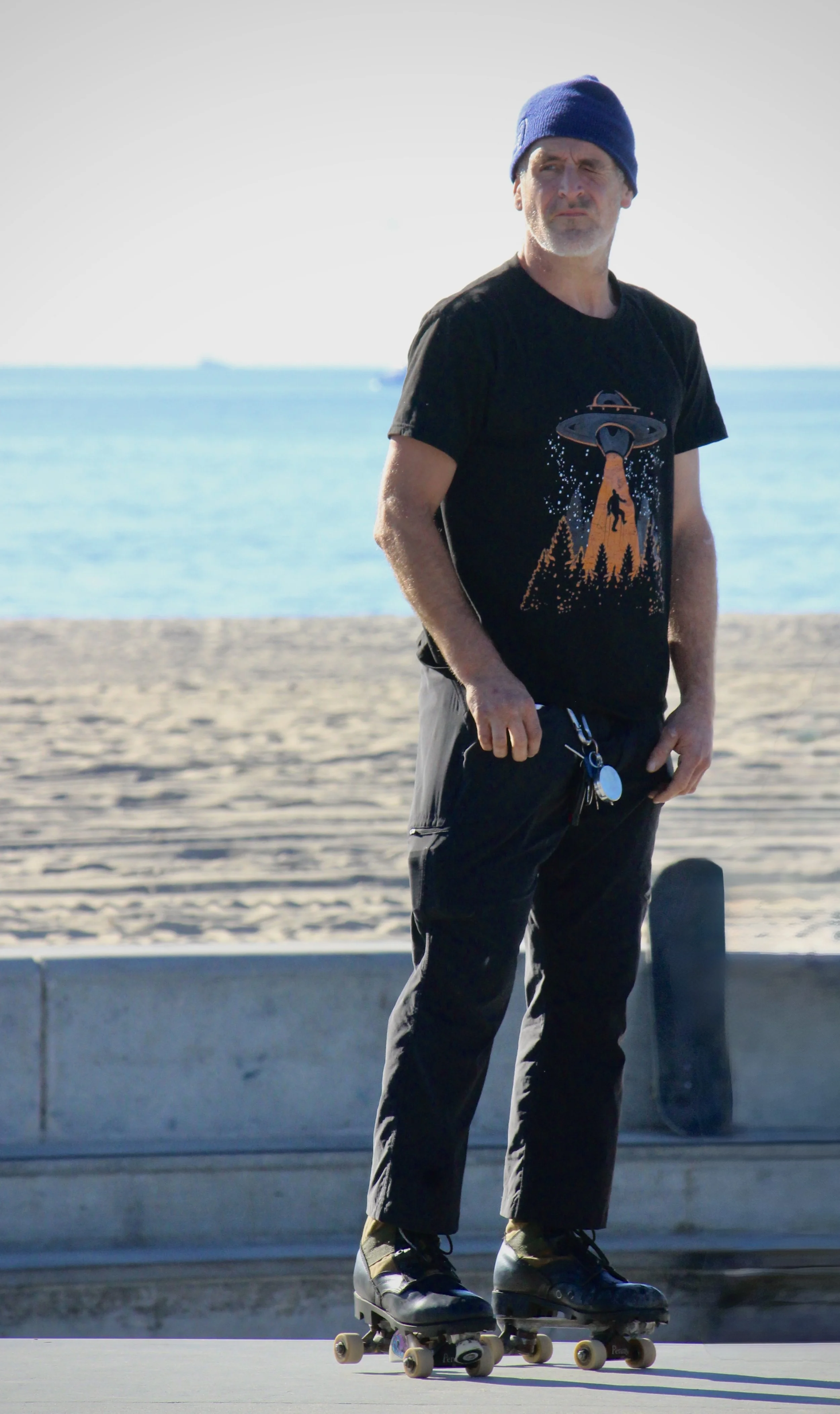 A man wearing a blue beanie, black t-shirt with a UFO graphic, black pants, and skate shoes roller skating outdoors by the water.
