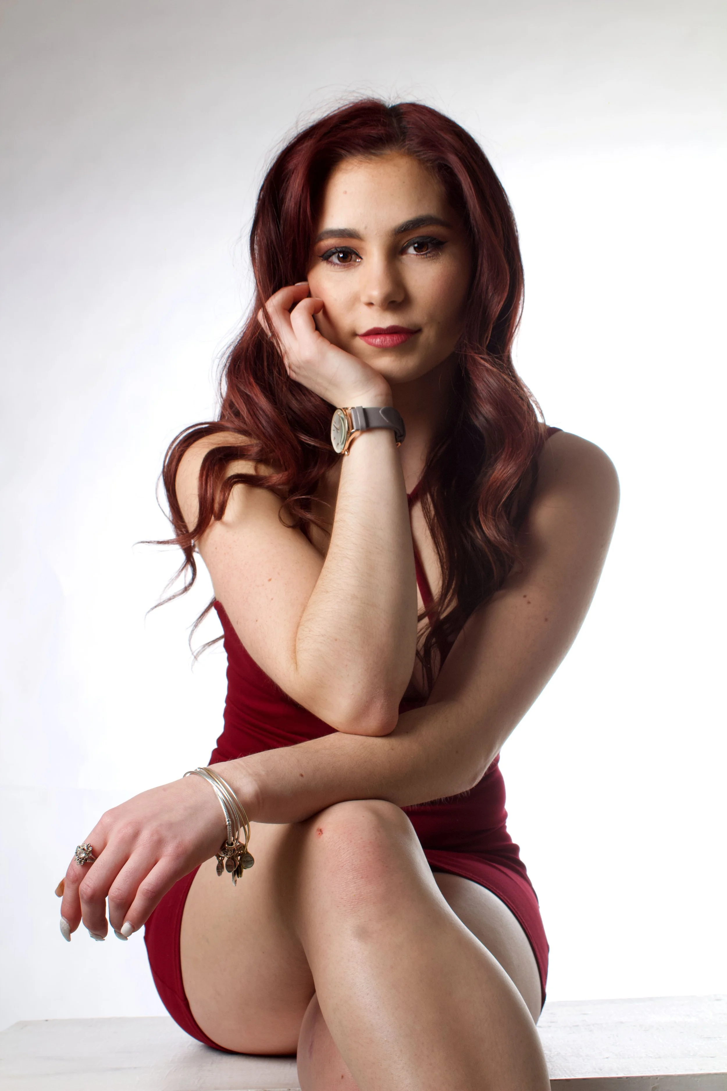 A young woman with long, wavy red hair and light skin sitting on a white bench against a plain white background. She is wearing a sleeveless, deep red dress, jewelry including bracelets and rings, and a watch. She has a confident expression with her 