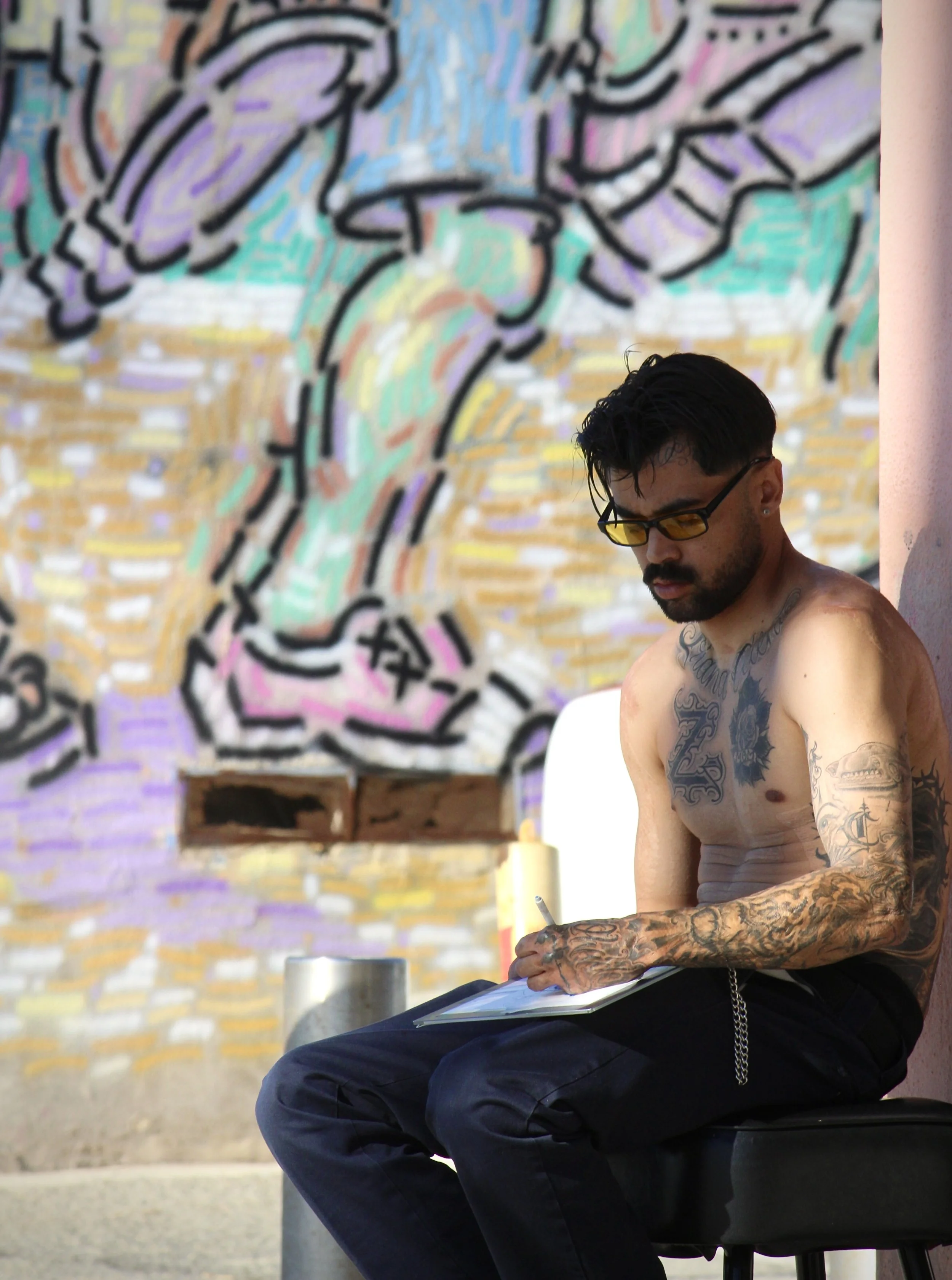 A shirtless man with tattoos, black hair, and glasses sitting outdoors, writing or drawing in a notebook, with a colorful mural in the background.