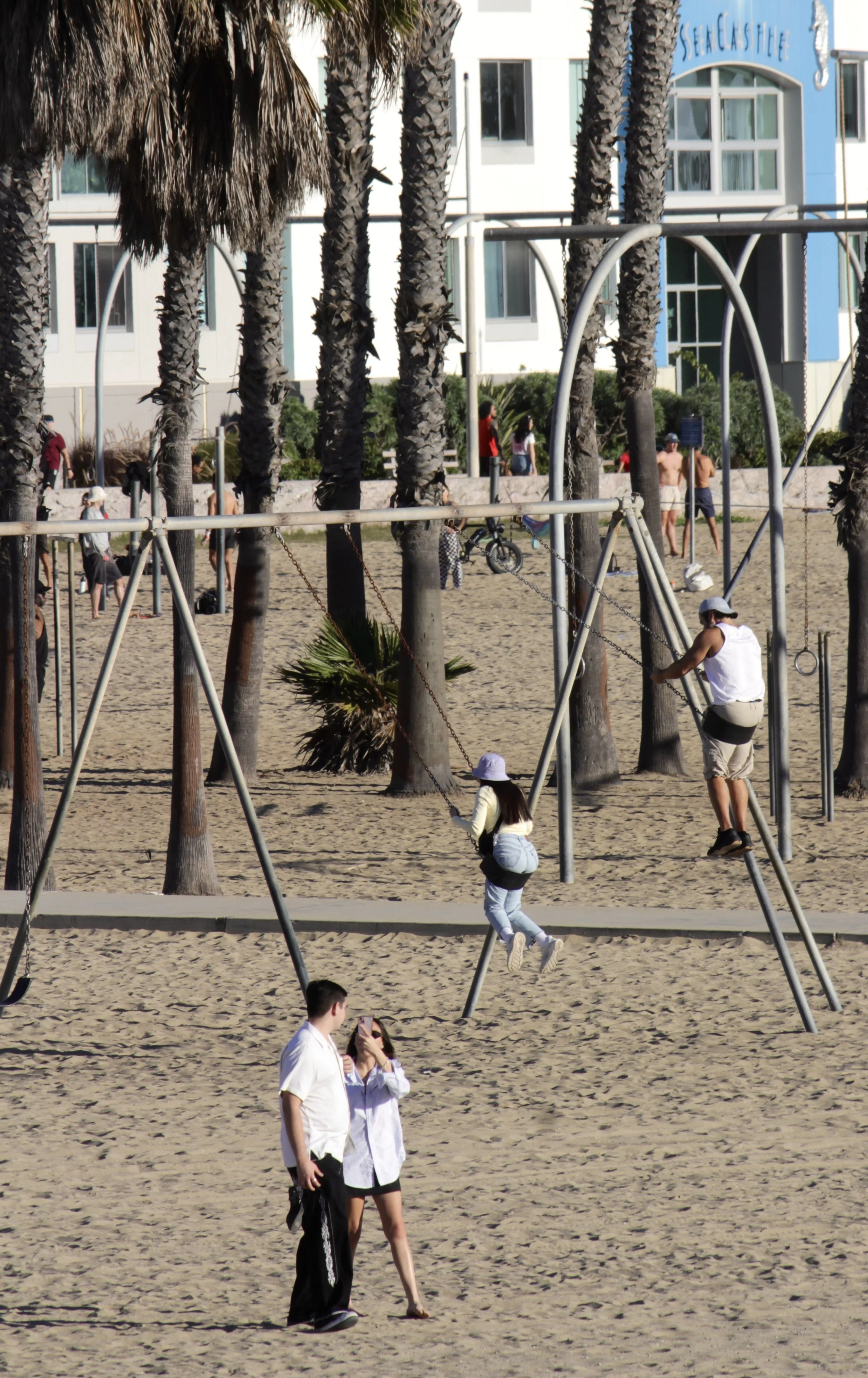 Beach playground with swings, palm trees, and people walking and playing on the sand, some in swimwear and casual clothing, with a blue and white building in the background.