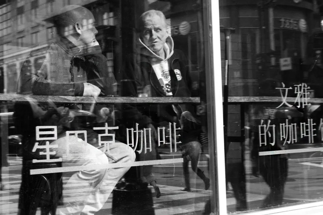 Two men sitting inside a café, seen through its glass window with reflections of the street and pedestrians outside.