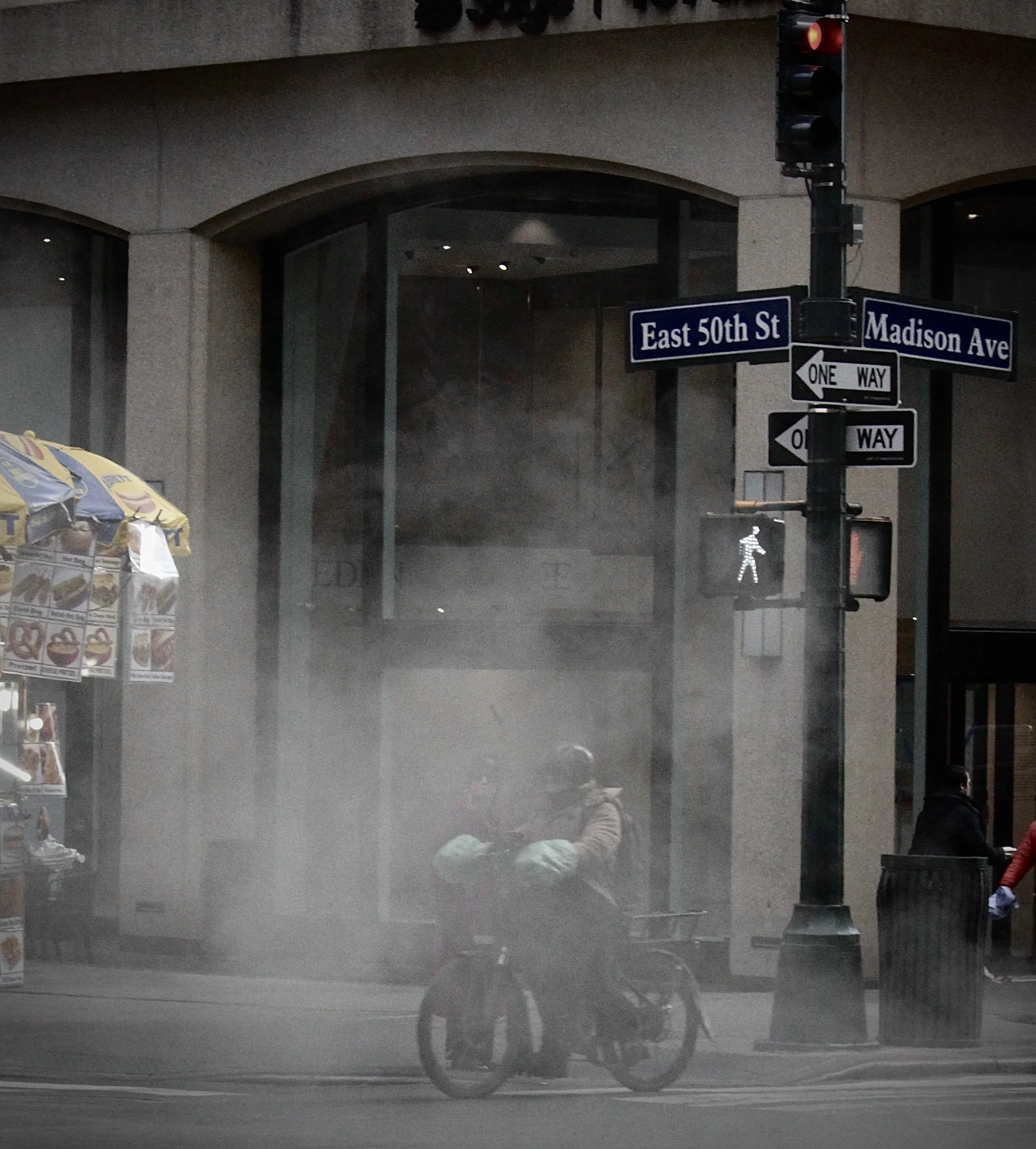 Street corner with traffic light showing red, street signs for East 50th Street and Madison Avenue, a pedestrian crossing signal, and a part of a storefront with advertisements for ice cream and other snacks visible on the left side.