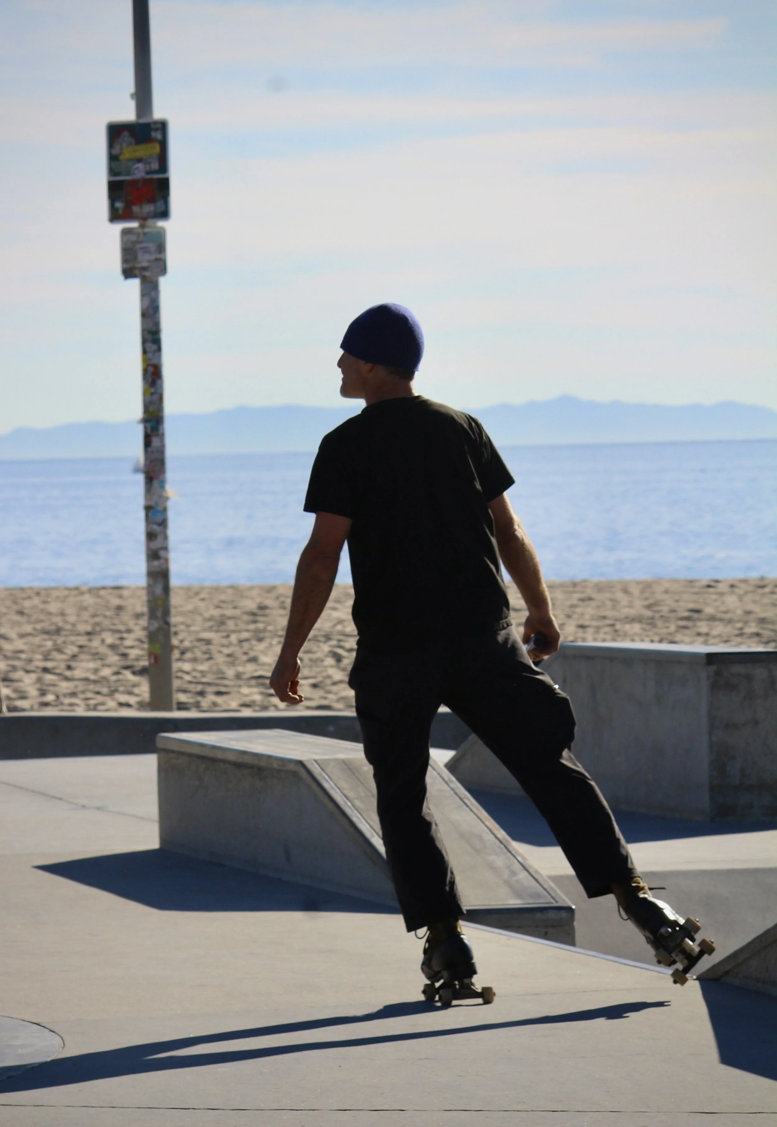 A person wearing a purple beanie and black clothing roller skating at a skate park near the beach, with mountains in the background.