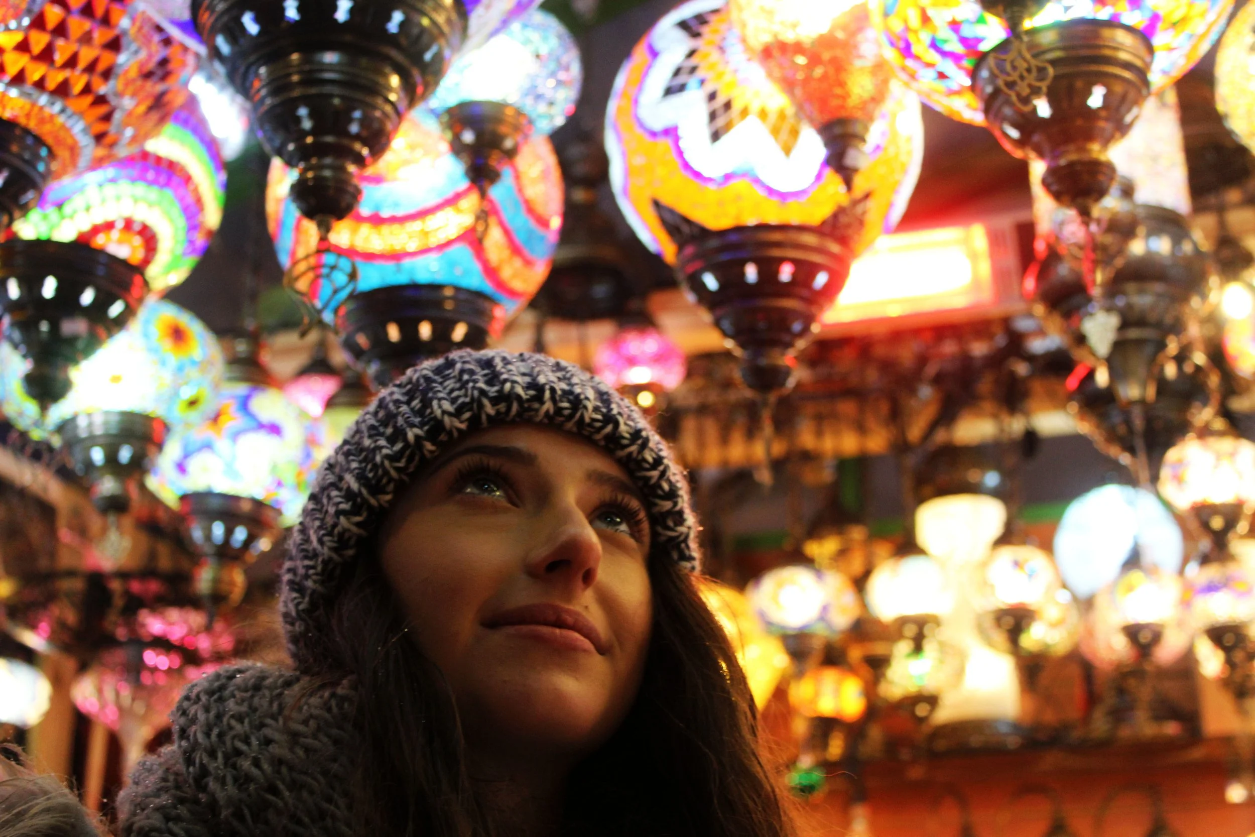 Close-up of a young girl with a knit hat, looking upward at colorful, illuminated mosaic glass lanterns hanging from the ceiling.
