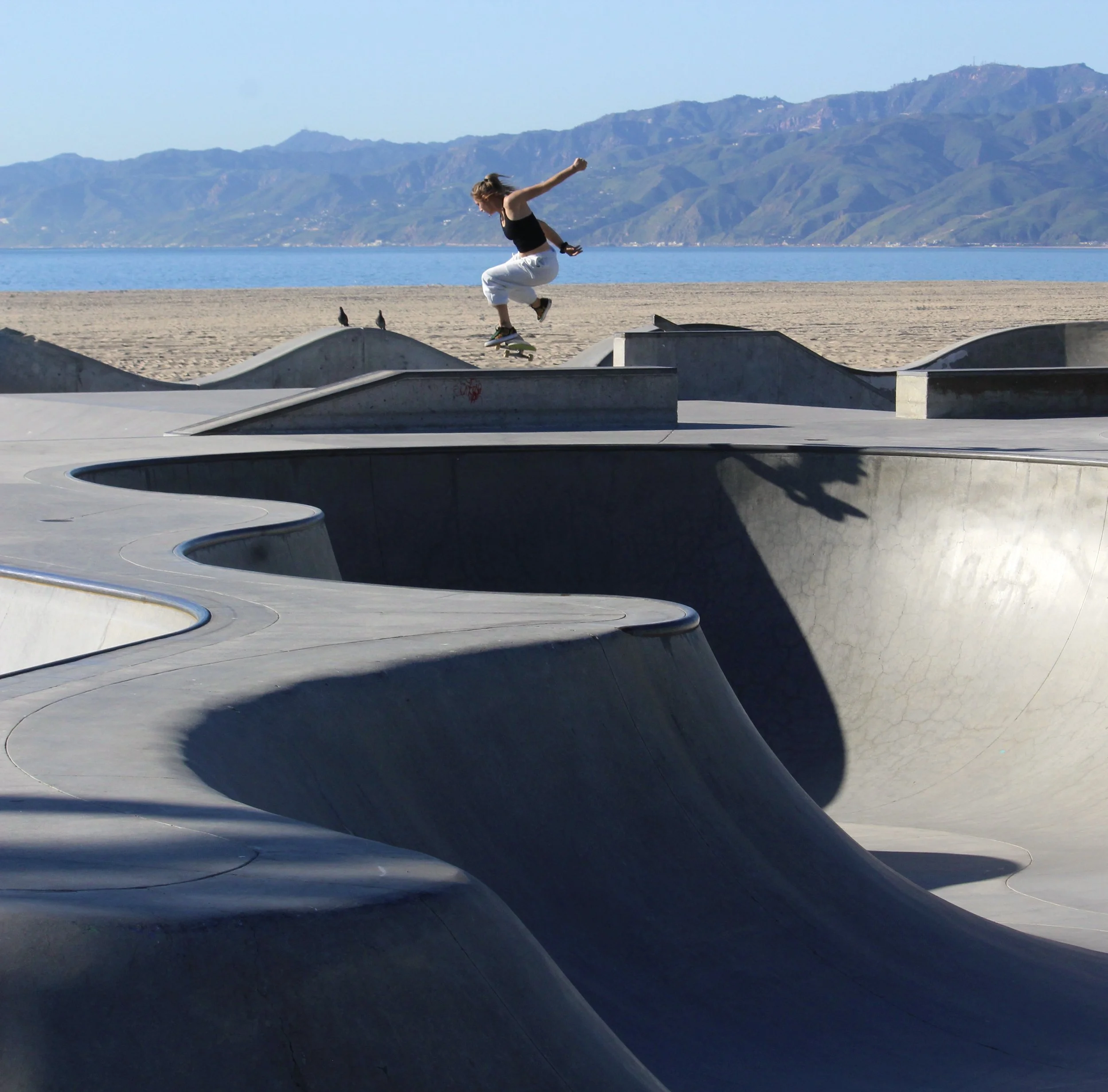 A person skateboard tricking on concrete ramps near a beach with mountains and water in the background.