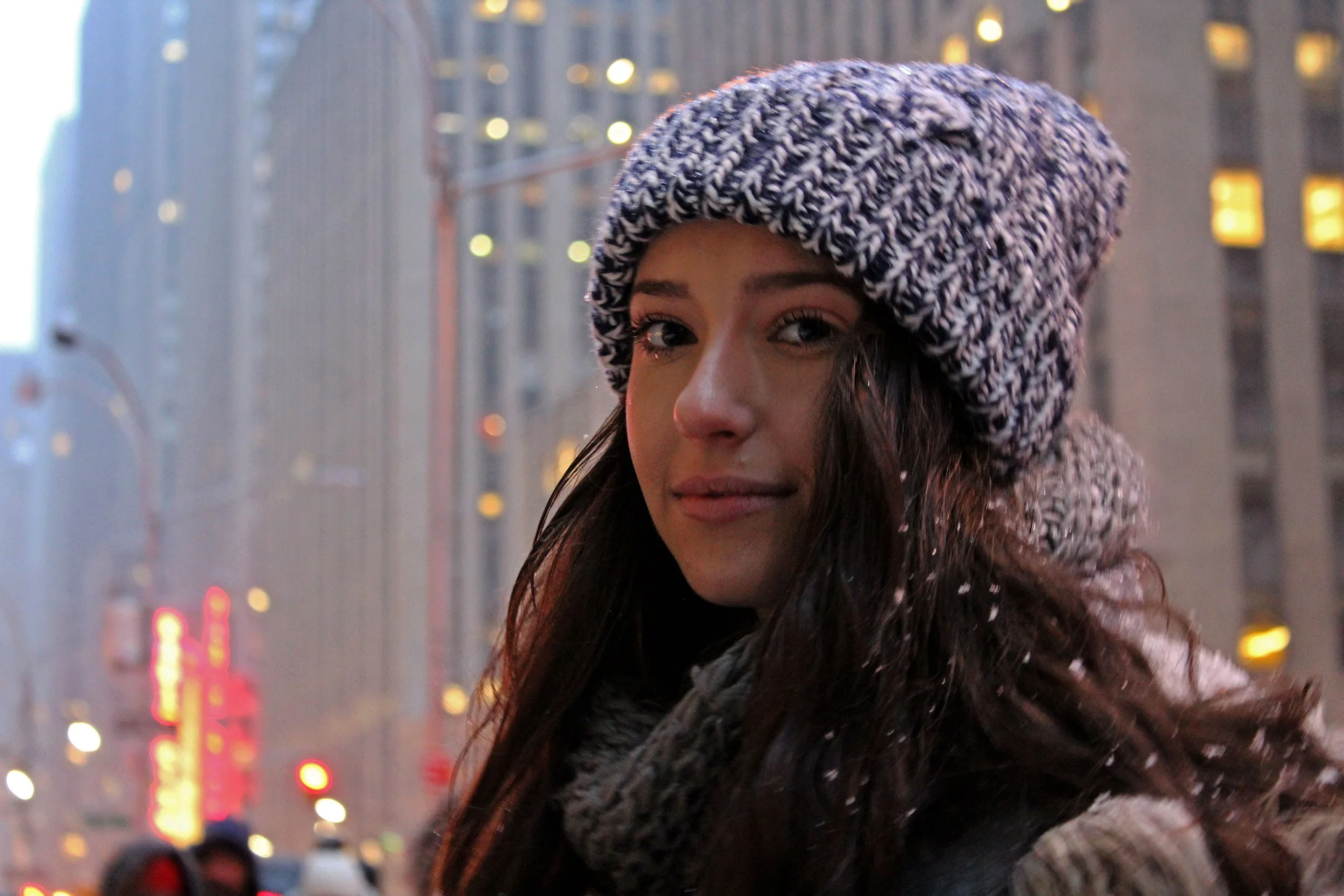 A young woman with long dark hair smiling, wearing a chunky black and white knit beanie hat and a matching knit scarf. The background shows a cityscape with tall buildings and blurred city lights at dusk.