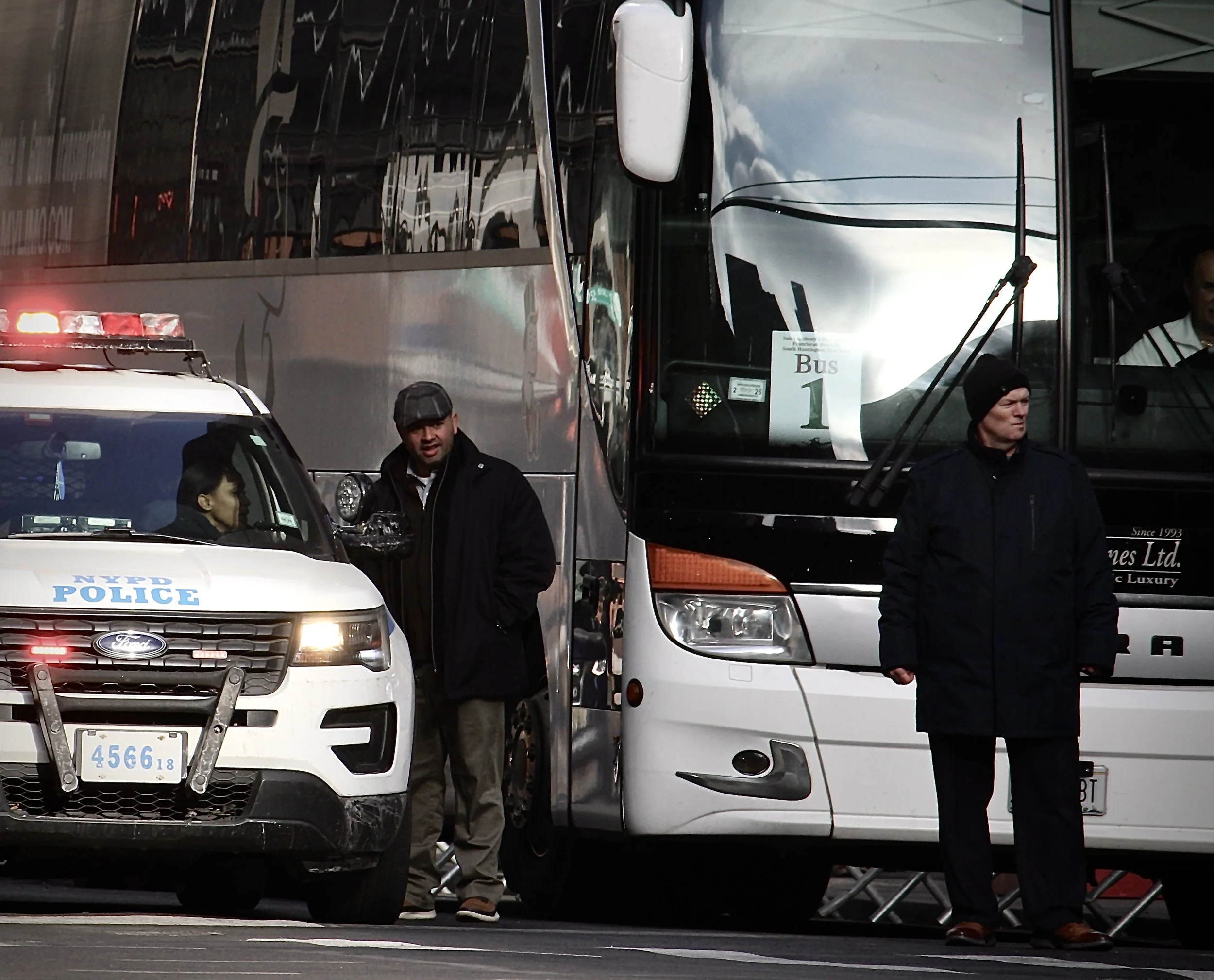 People standing near white and black buses and police vehicle at an outdoor location.