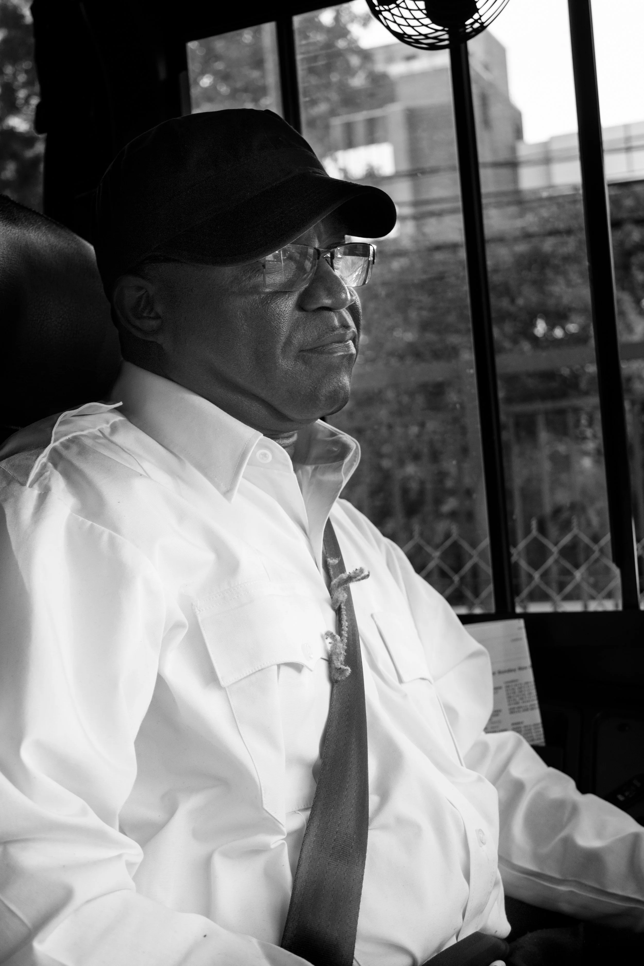 A man wearing glasses, a cap, and a white shirt sits with his eyes closed inside a vehicle or bus, with a window and some buildings visible in the background.