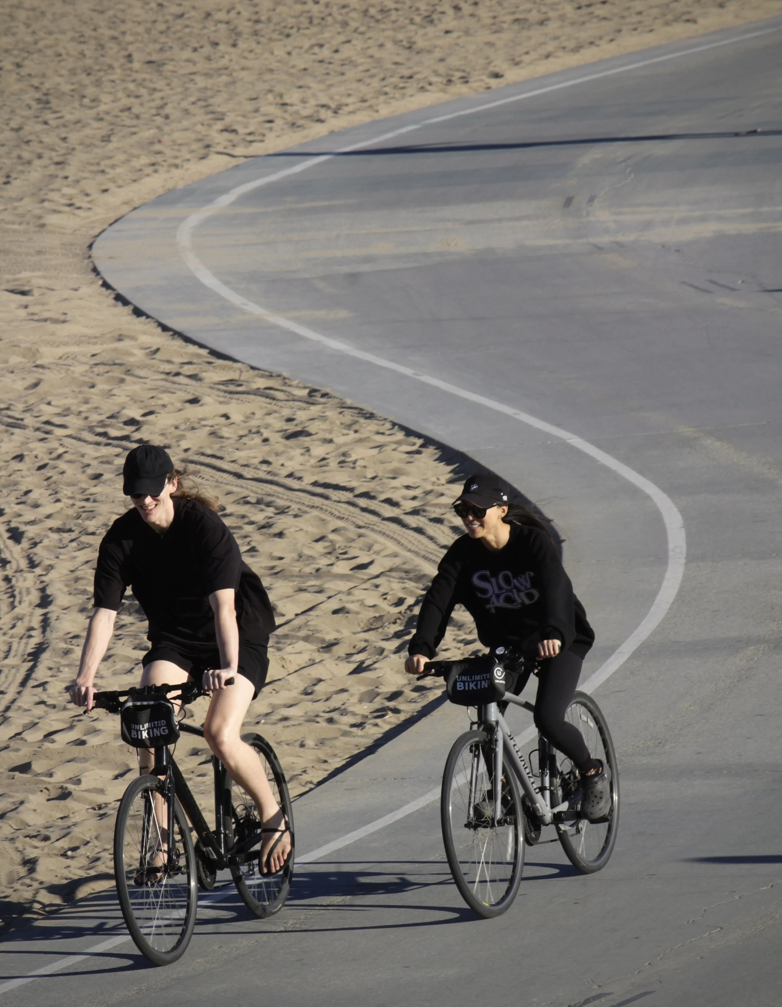 Two women riding bicycles on a paved beachside pathway, with sandy beach and sand dunes visible in the background, both smiling and wearing black clothing and sunglasses.