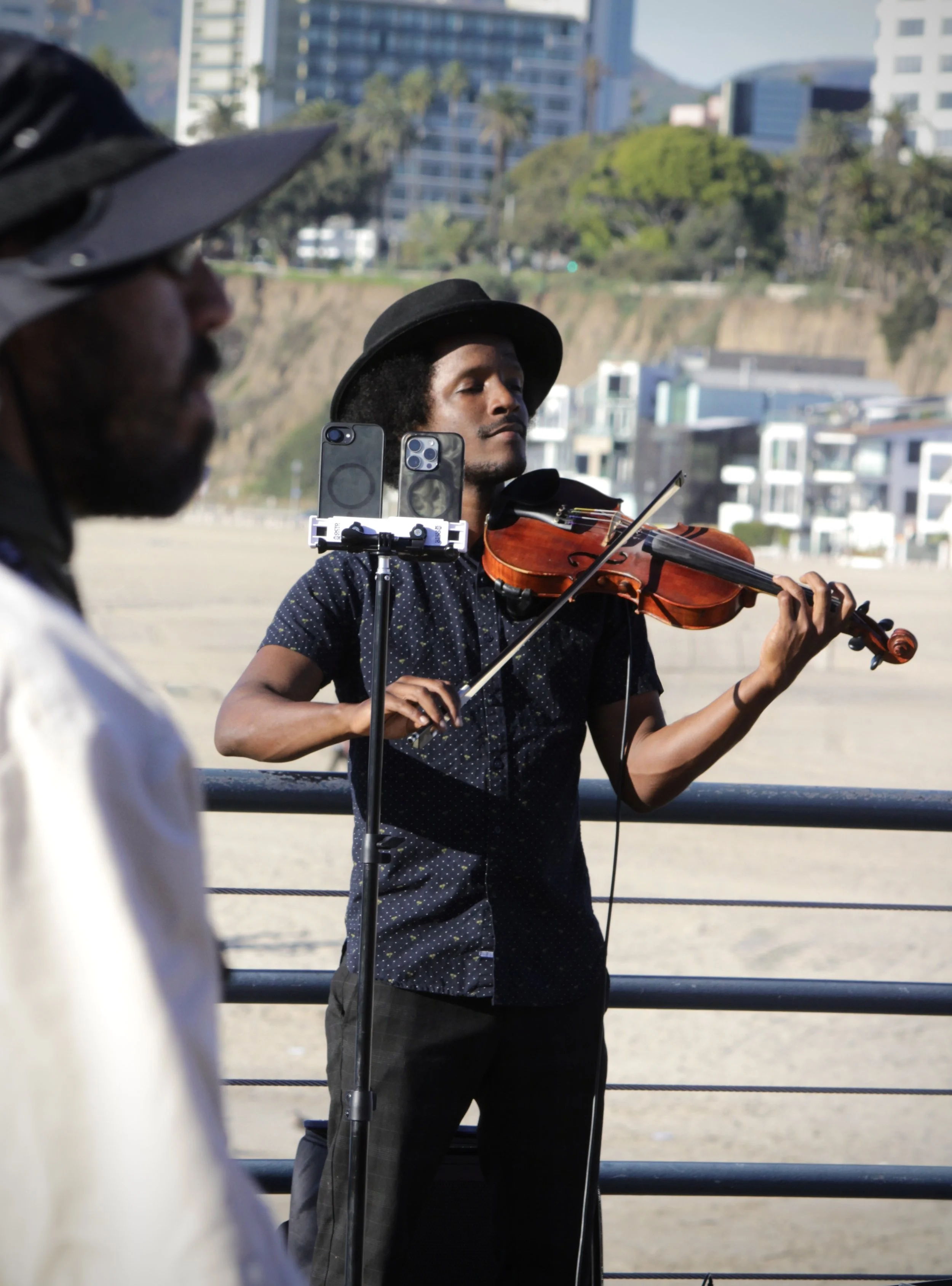A man playing the violin outdoors, using a smartphone stand to hold two phones, with a cityscape of tall buildings and trees in the background.