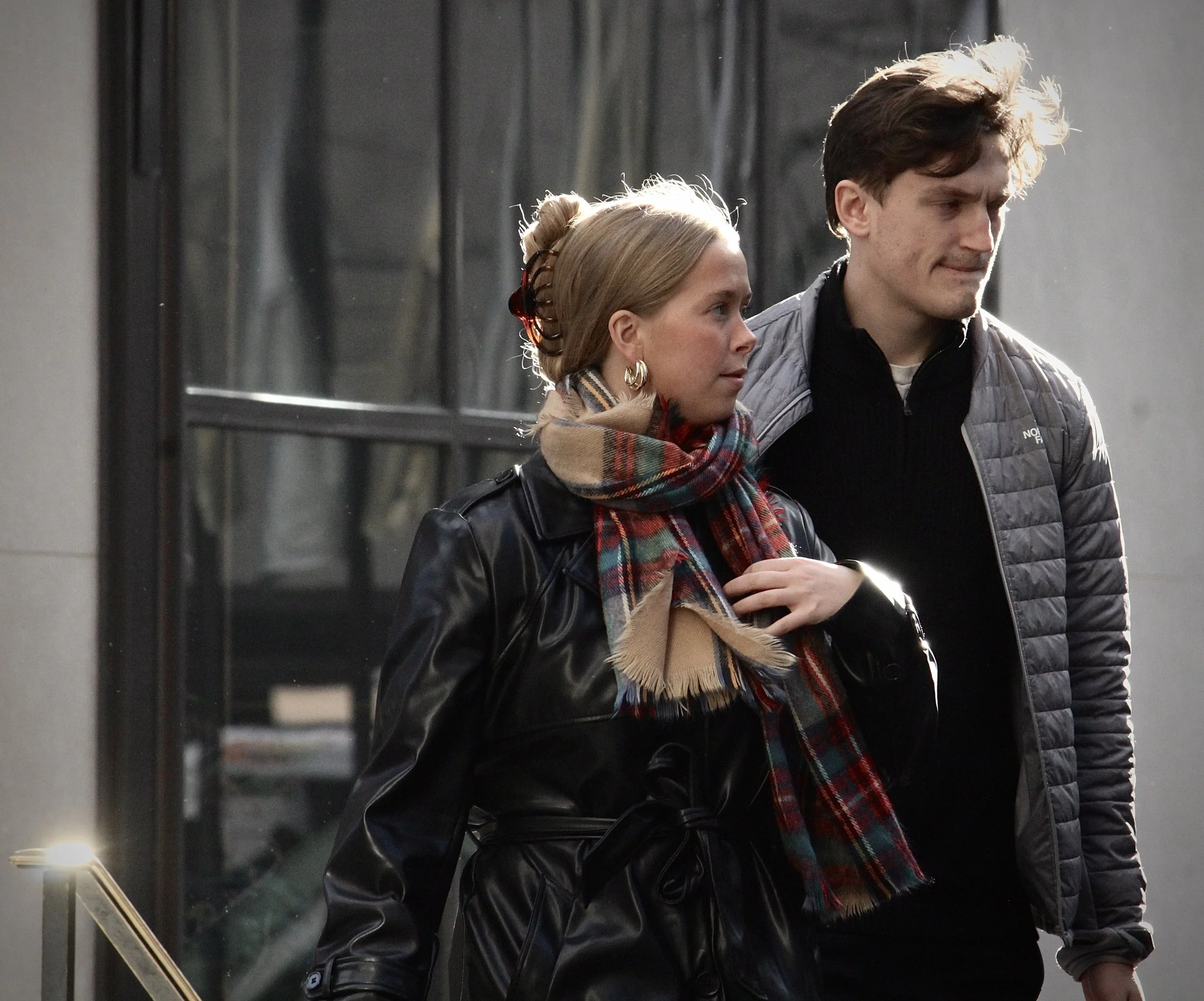 Two people walking outdoors beside a glass building, one man and one woman, both with serious expressions, the woman with blonde hair in a bun, wearing earrings and a plaid scarf, the man with brown hair, wearing a gray quilted jacket.