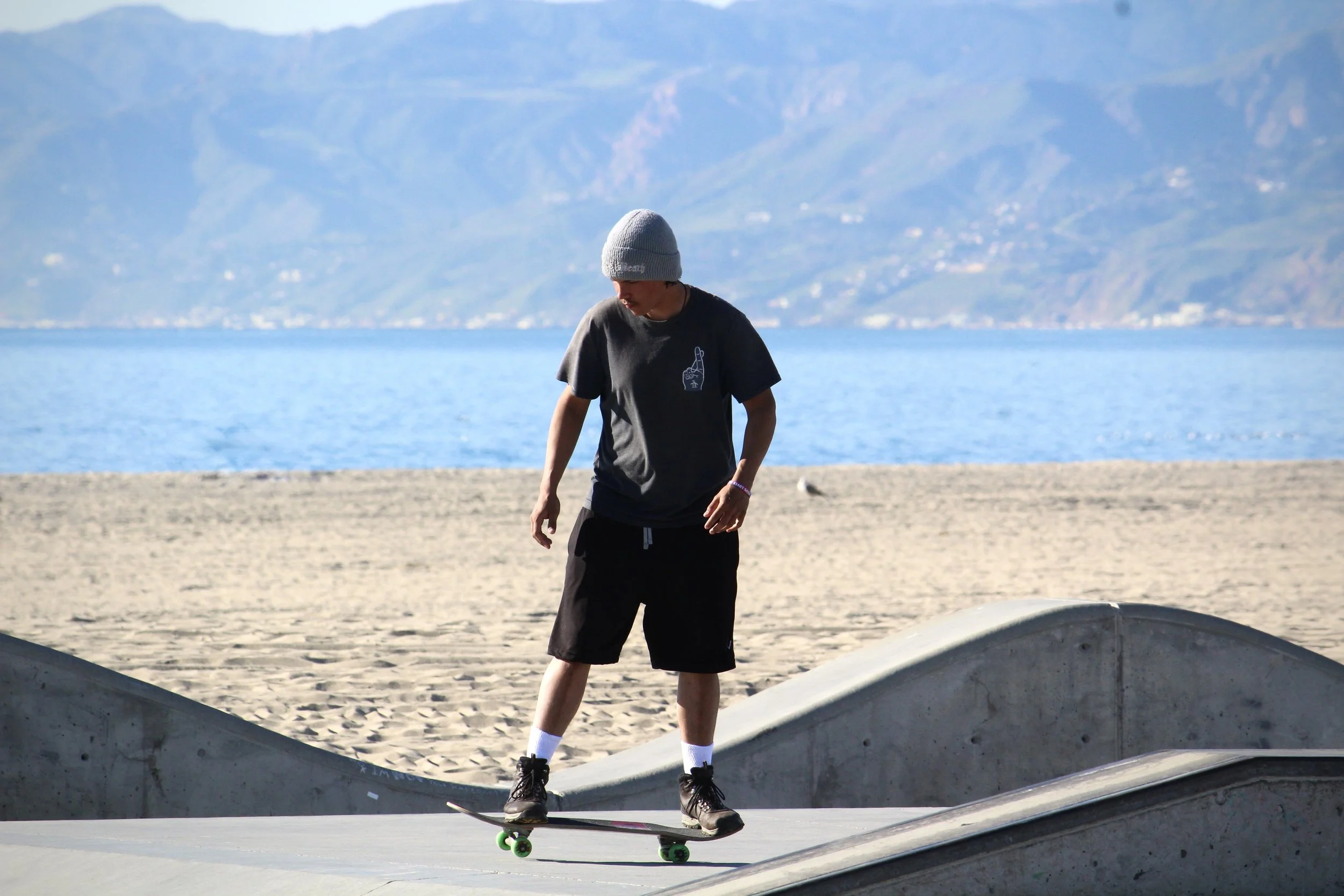 A young person wearing a gray beanie, black t-shirt with a hand gesture on it, and black shorts riding a skateboard at a skate park near a body of water with mountains in the background.