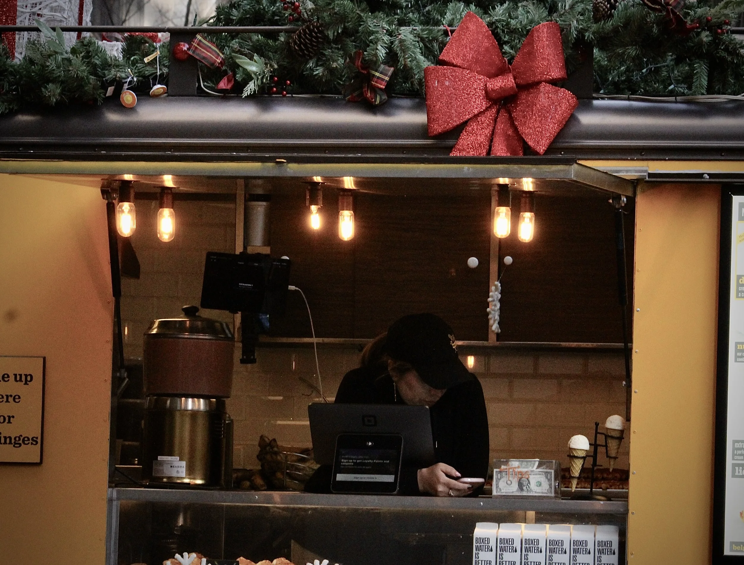 A decorated Christmas garland with red and green accents, including a large red glittery bow, hanging above a dark food kiosk. The interior of the kiosk has warm lighting, a person using a phone, and various items on the counter such as a cash regist