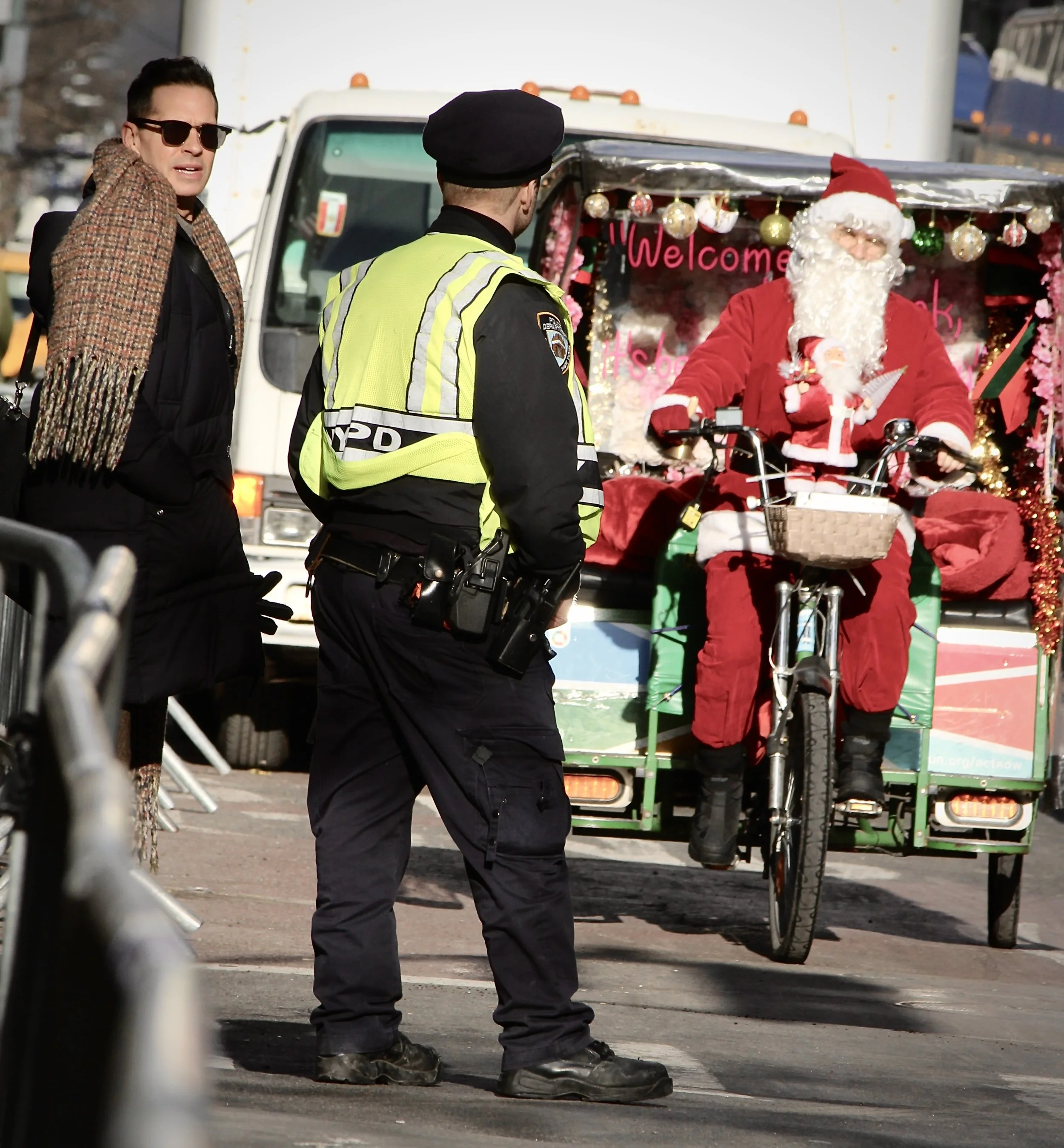A police officer and a man in sunglasses are having a conversation near a truck decorated for Christmas, with a Santa Claus figure on a bicycle in front of it.