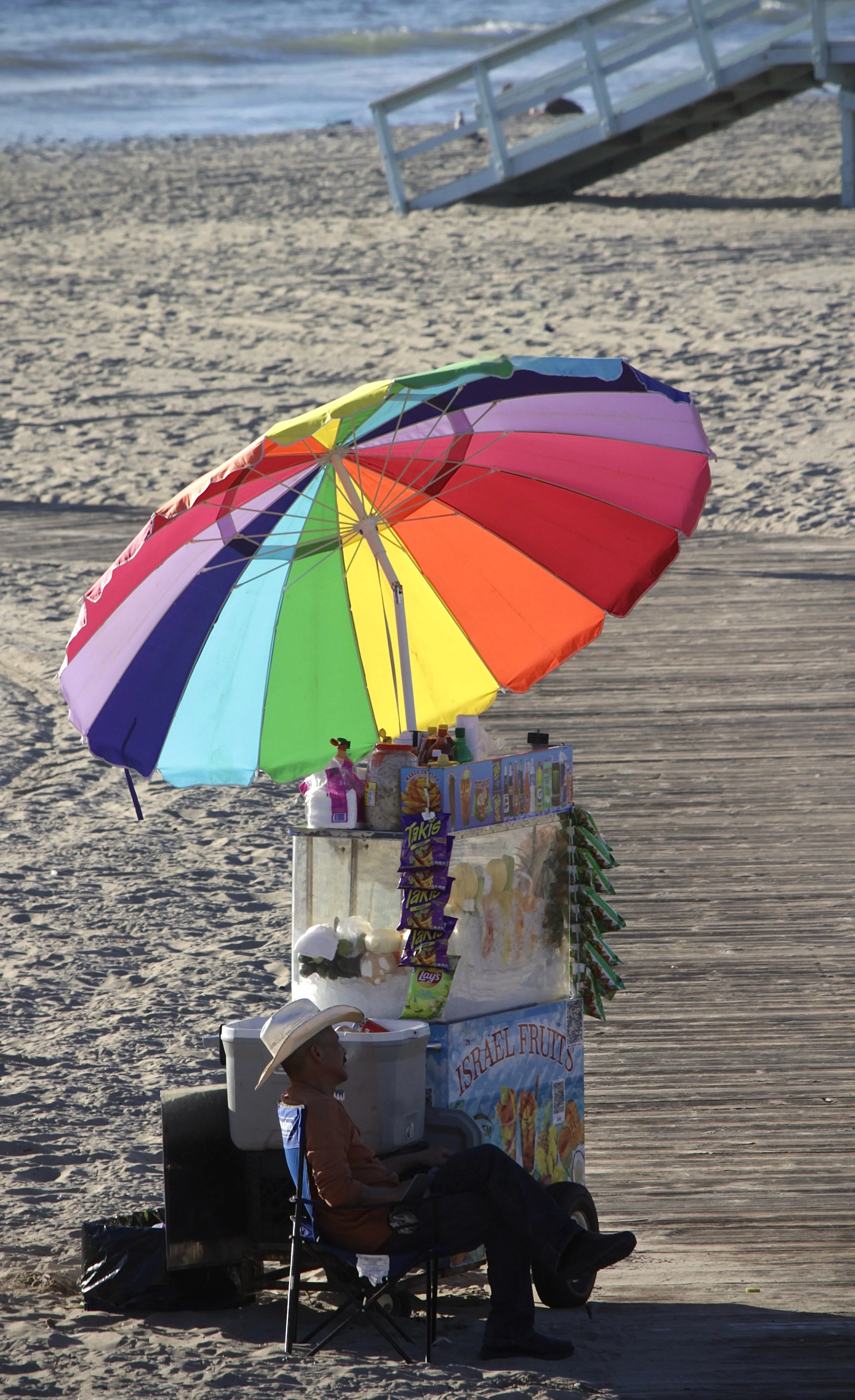 A man sitting in a chair under a large rainbow-colored umbrella on a sandy beach next to a snack stand selling fruit and snacks.