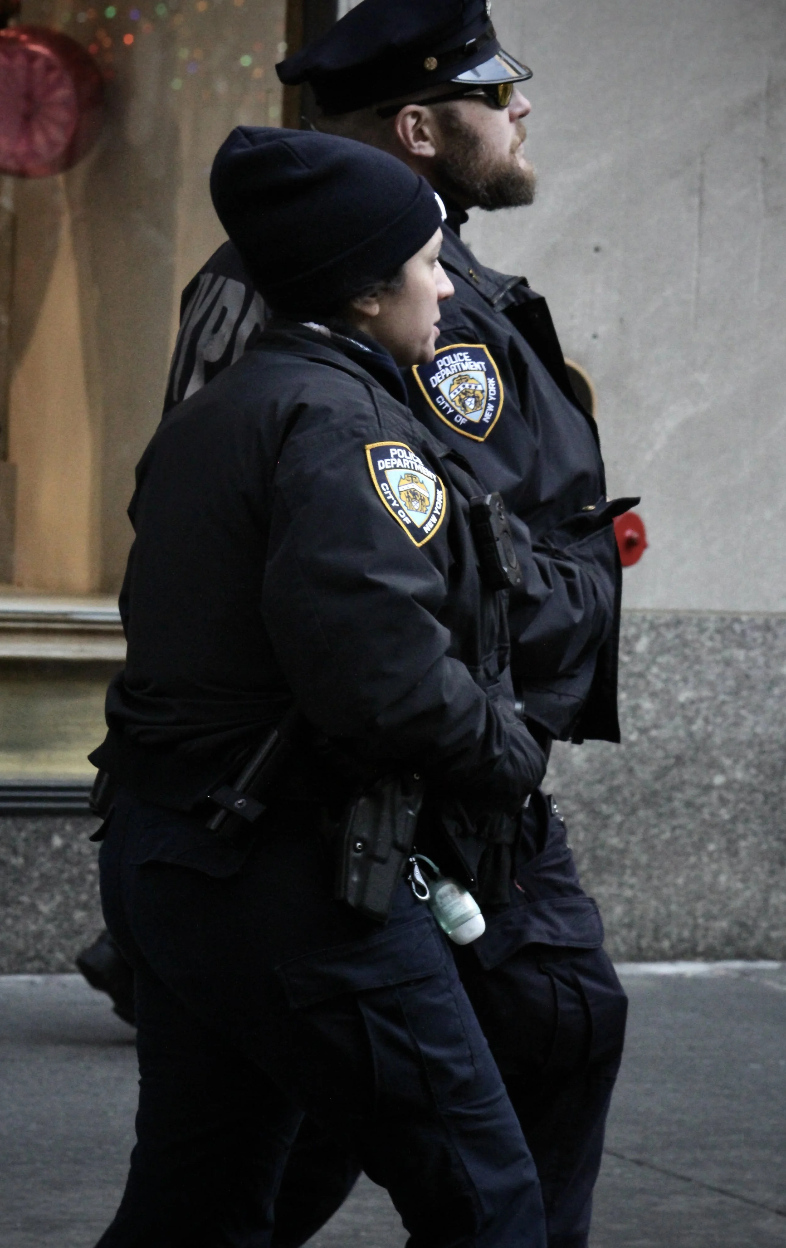 Two police officers standing side by side, wearing dark uniforms with police department patches, caps, and tactical gear, walking along a city sidewalk with a concrete wall in the background.