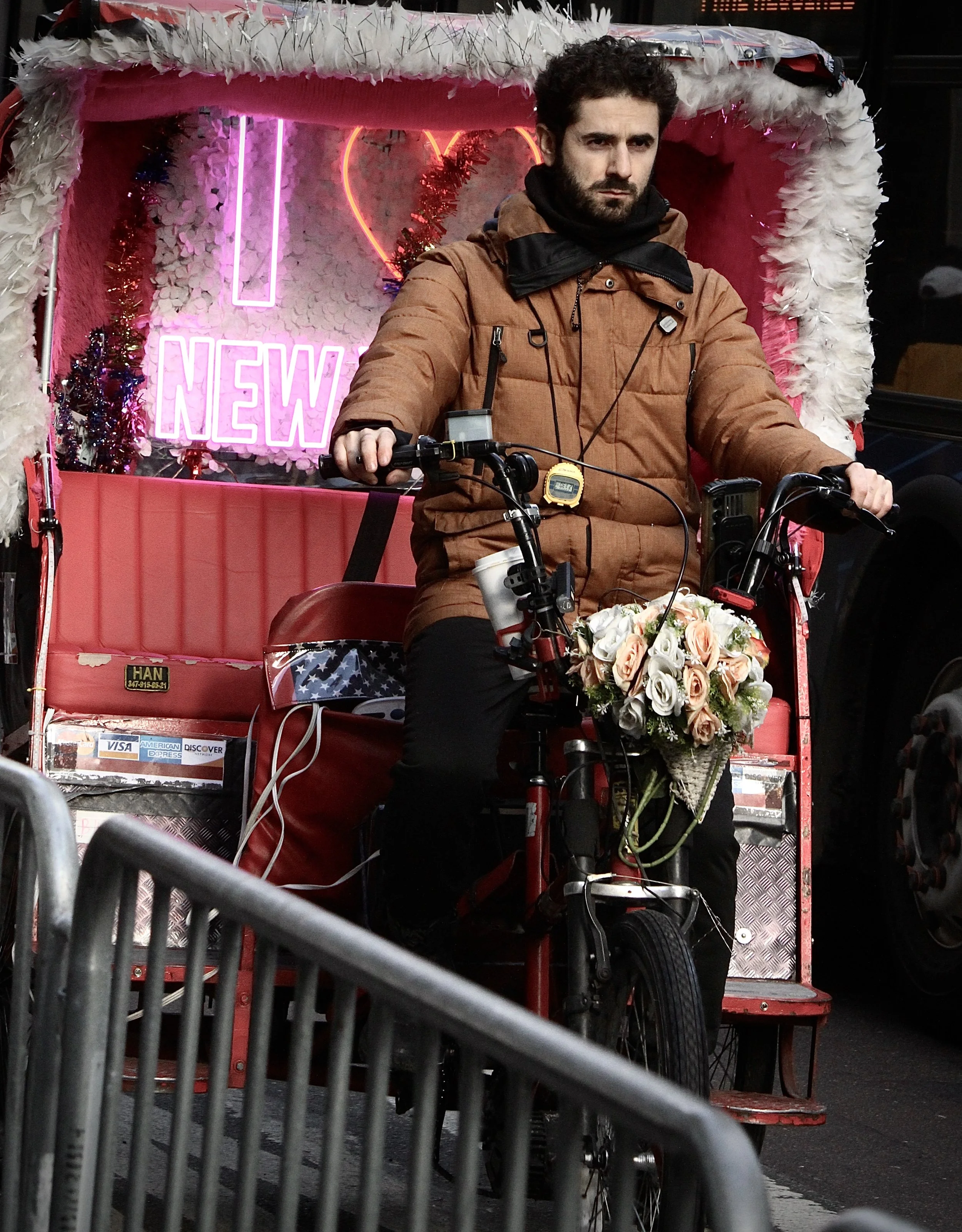 A man with a beard and curly hair riding a decorated pedicab with a bouquet of flowers on the front. The background features pink and red neon signs, including one that says 'NEW' inside a heart shape, and pink and white floral and feather decoration
