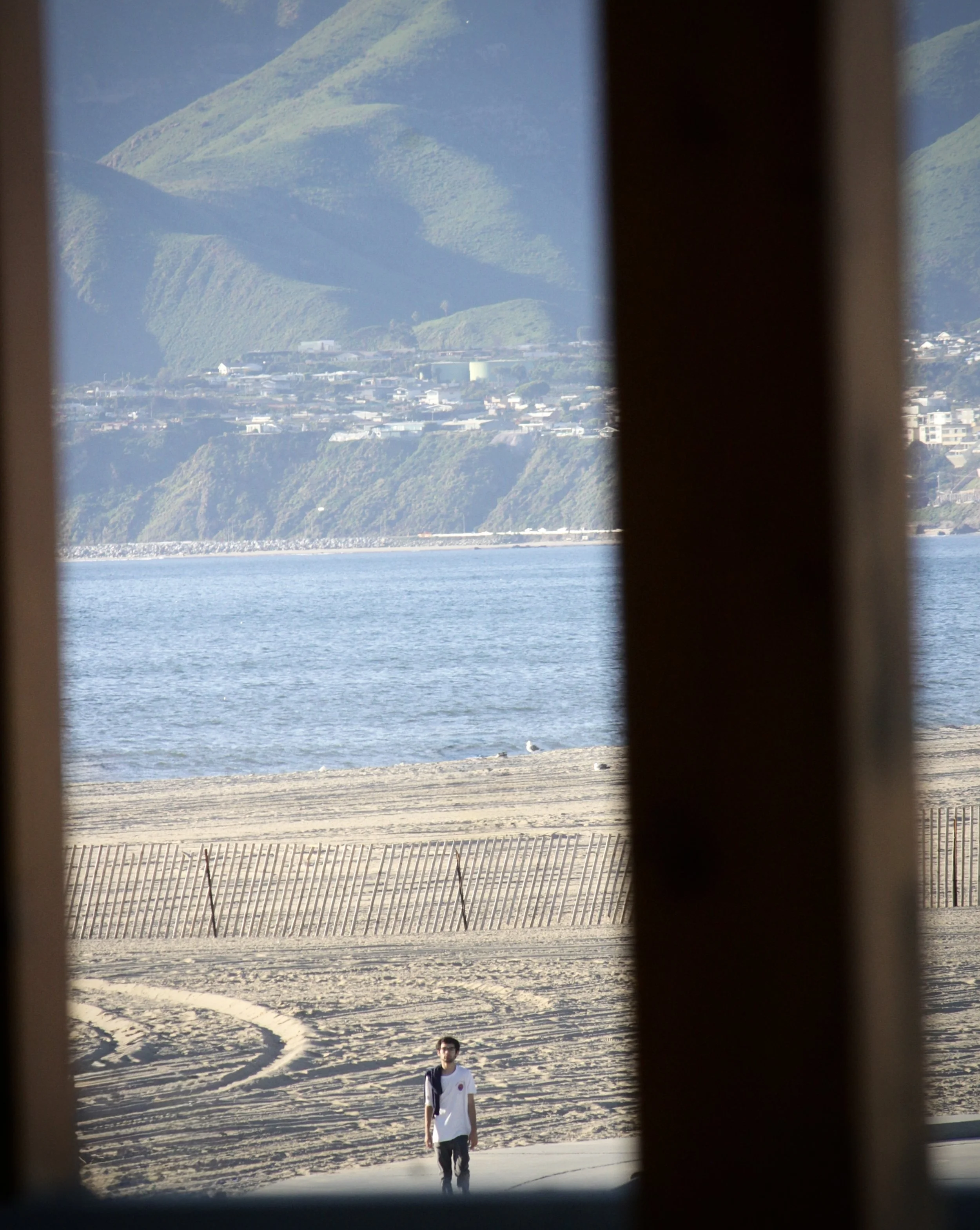 A person walking on the beach beach with hills and water in the background, taken through wooden slats.