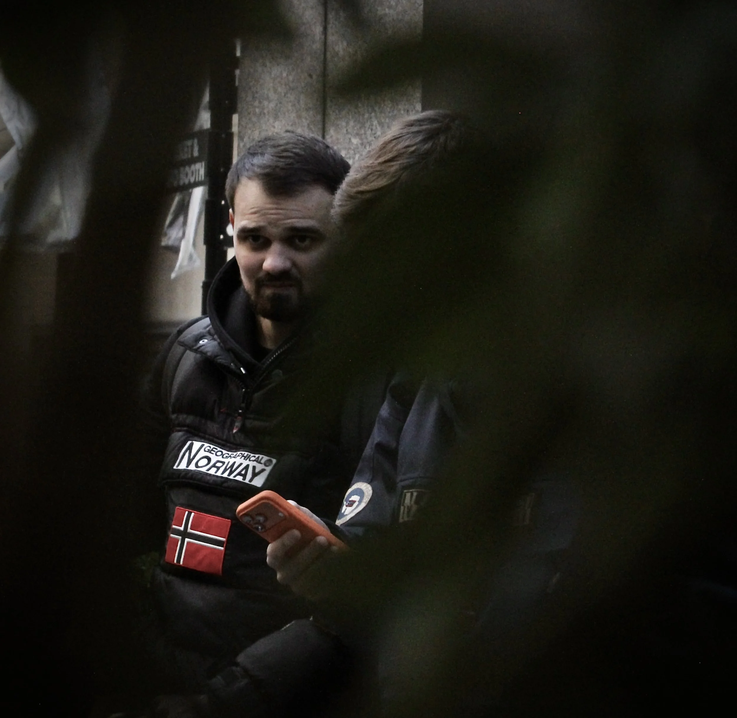 A man with dark hair and a beard, wearing a black jacket, is looking at his phone. The background suggests he is in a public or indoor space with a granite or stone wall behind him.