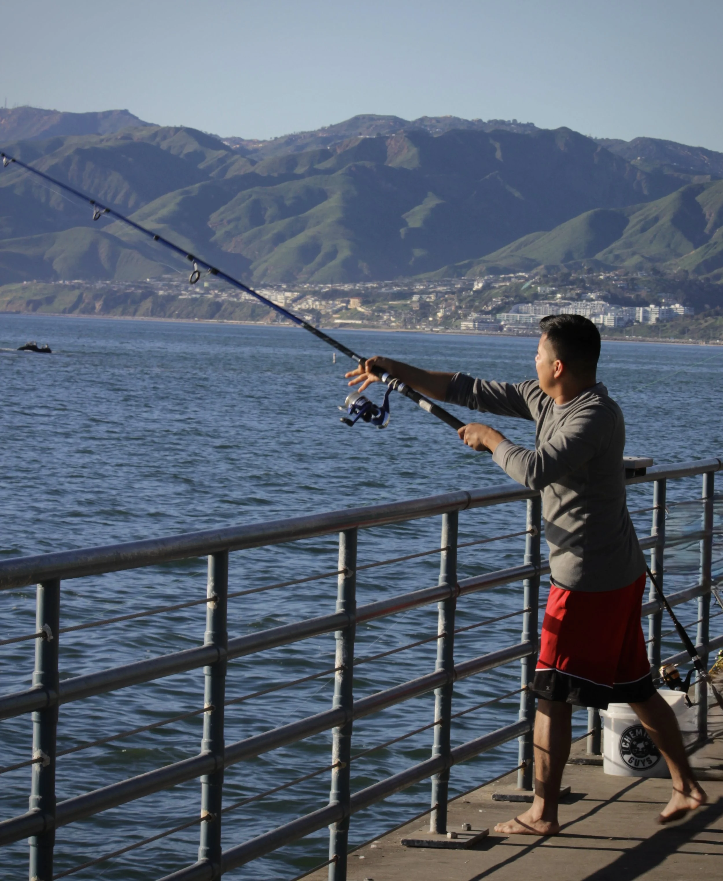 Young man fishing from a pier with mountains in the background, wearing shorts and a long-sleeved shirt, standing on a small scale on the pier, next to a bucket, overcast sky.