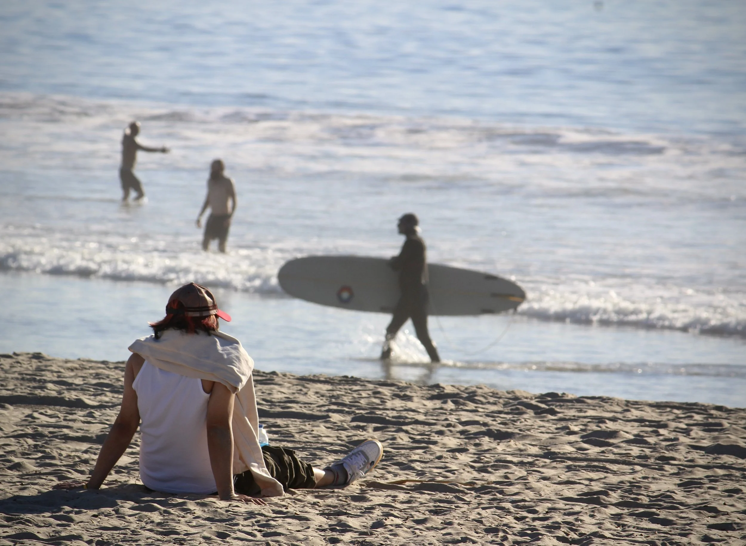 A young girl sitting on a sandy beach facing the water, wearing a white shirt and a red cap with a white stripe. In the background, people are enjoying the ocean, with one person surfing and others wading in the water.