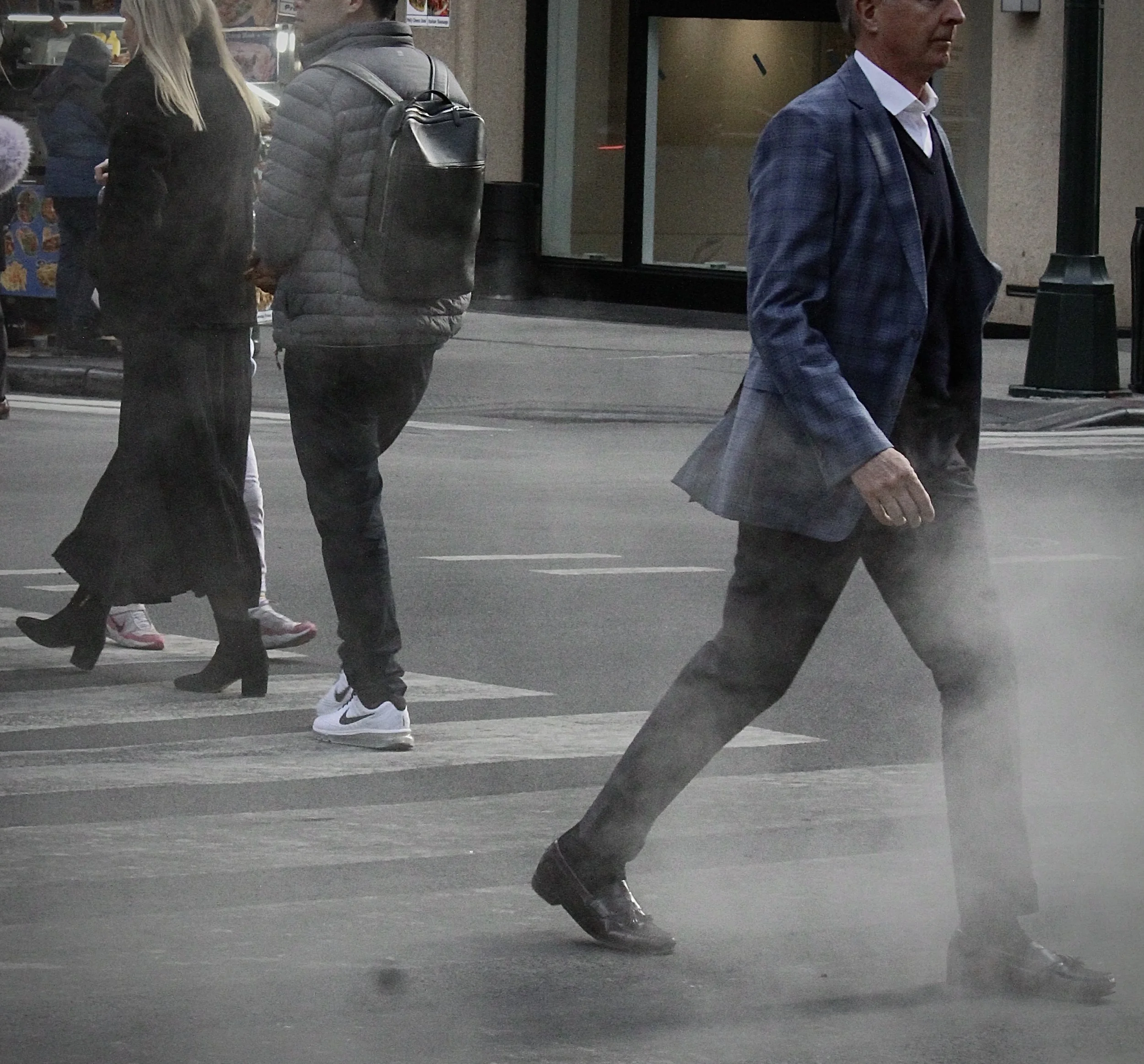People walking on a city sidewalk at dusk, dressed in urban clothing including jackets and sneakers, with store windows in the background.