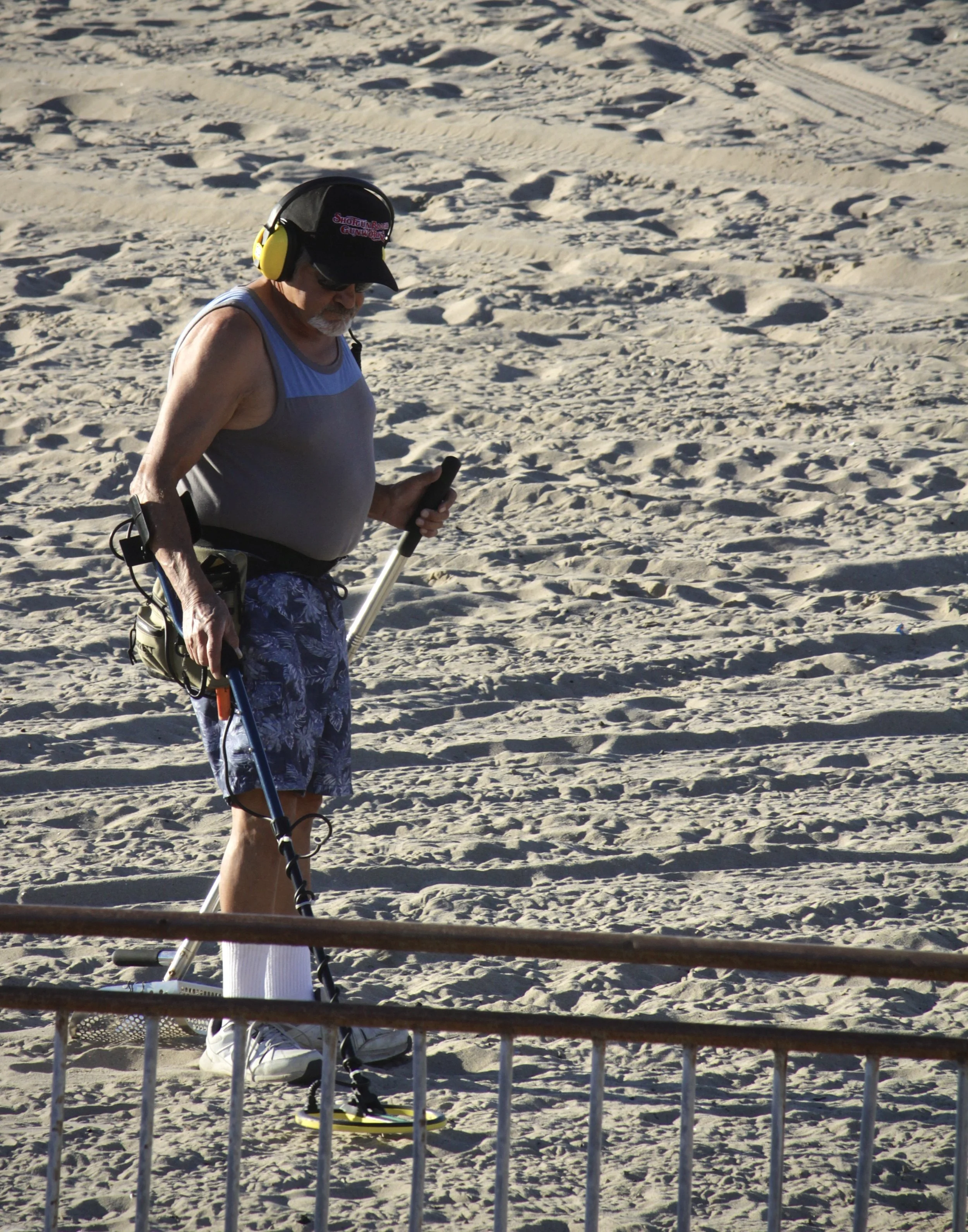 An elderly man on the beach with a metal detector, wearing a hat, sunglasses, and headphones, holding the metal detector's handle, with a picnic basket and other items beside him.