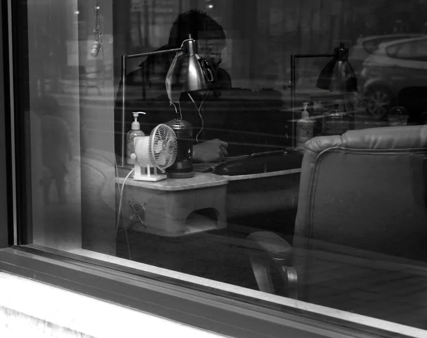 A person working at a desk, viewed through a window, with desk lamps, a small fan, and bottles on the desk inside a room.