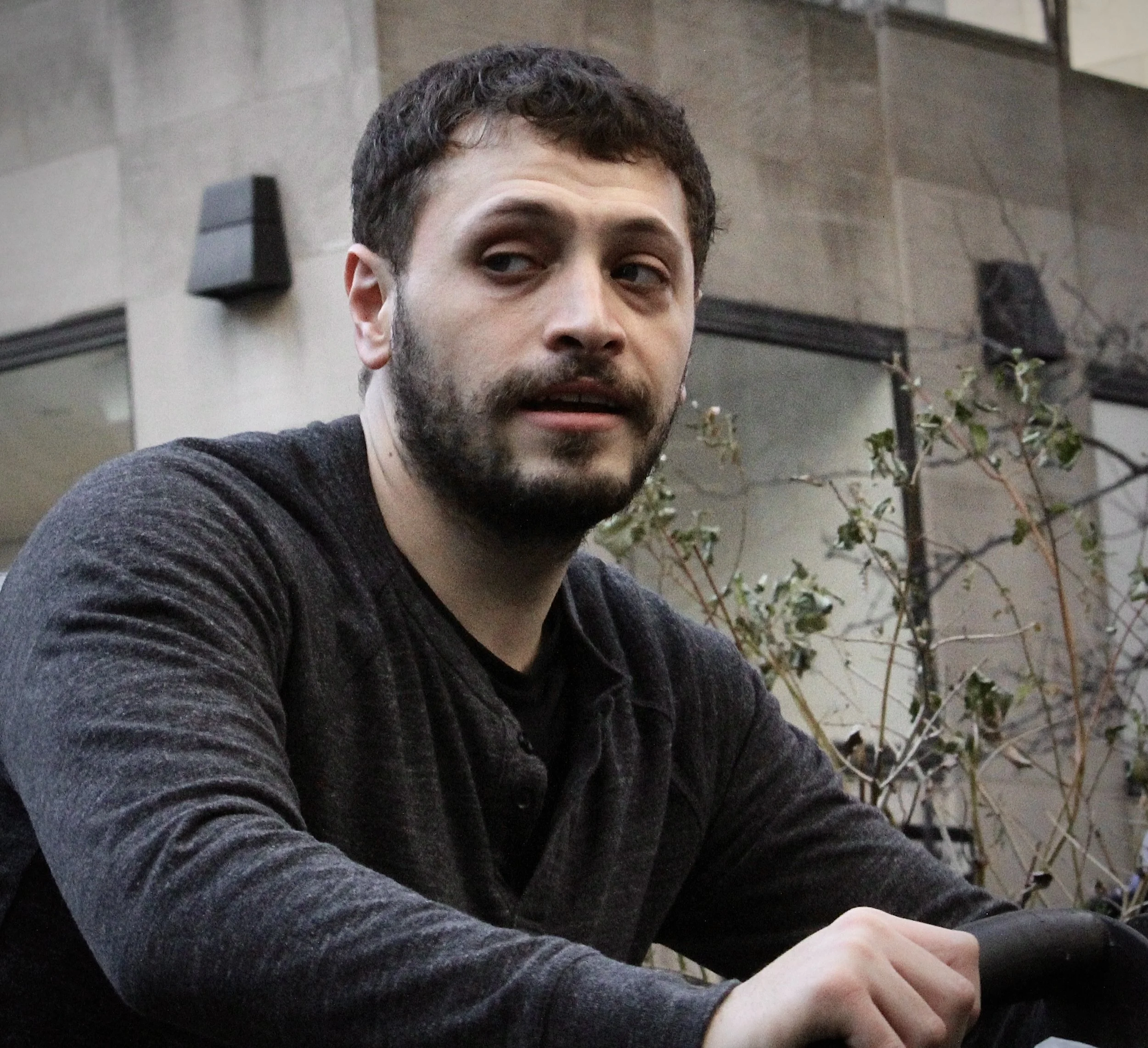 A man with dark, curly hair and a beard is looking to his right. He is wearing a dark gray, ribbed sweater. The background shows a beige wall with some black speakers, a branch with green leaves, and part of a white ceiling.