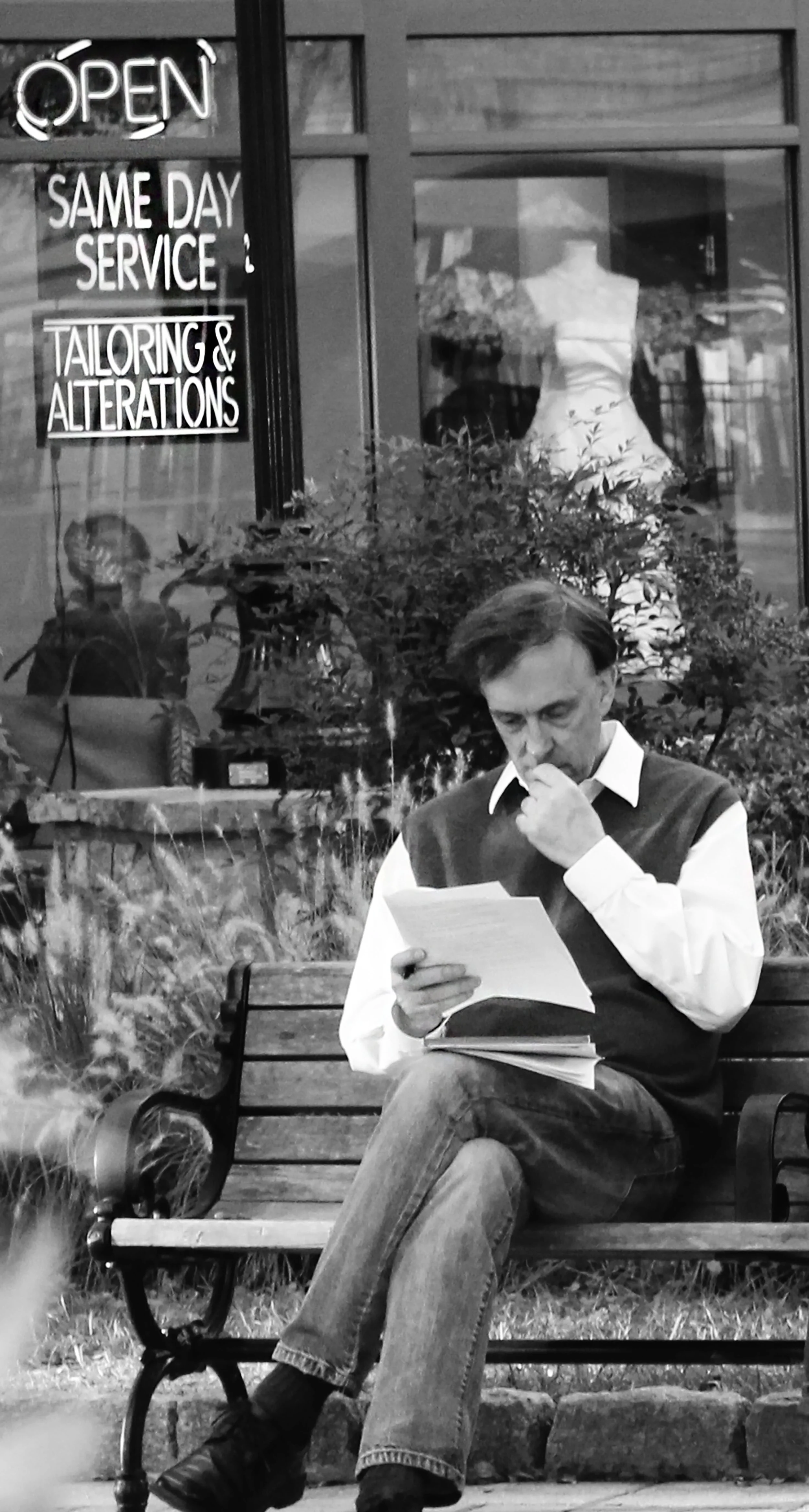 A man is sitting on a park bench reading a document, with left leg crossed over the right, in front of a shop window displaying a dress and signs that say "Open," "Same Day Service," and "Tailoring & Alterations."
