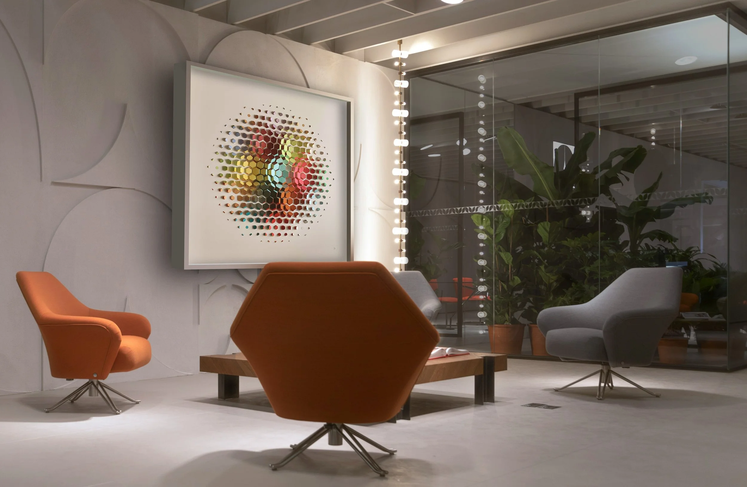 Modern office lounge area with colorful chairs, a wooden coffee table, large wall art with circular pattern, and indoor plants behind glass walls.