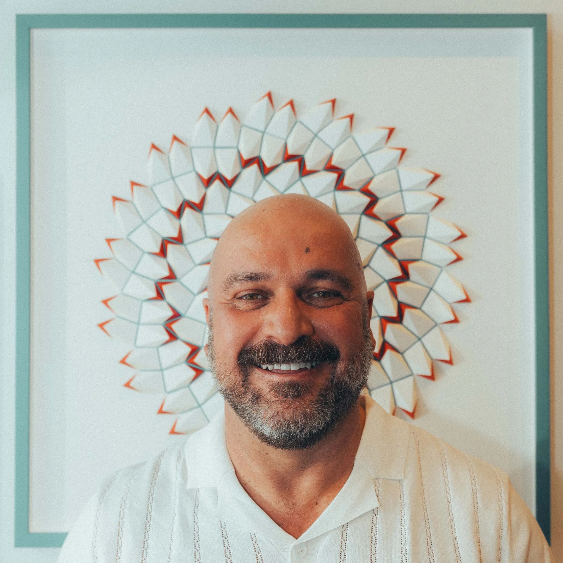 A smiling man with a beard and bald head poses in front of a decorative wall art piece.