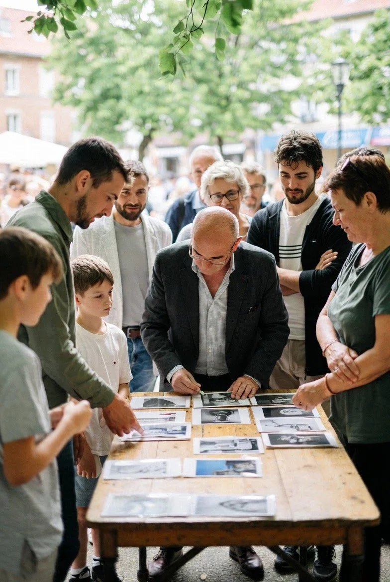 Un groupe de personnes, y compris des enfants et un homme âgé, qui regarde des photos sur une table en plein air. Le fond montre des arbres et une foule de personnes.