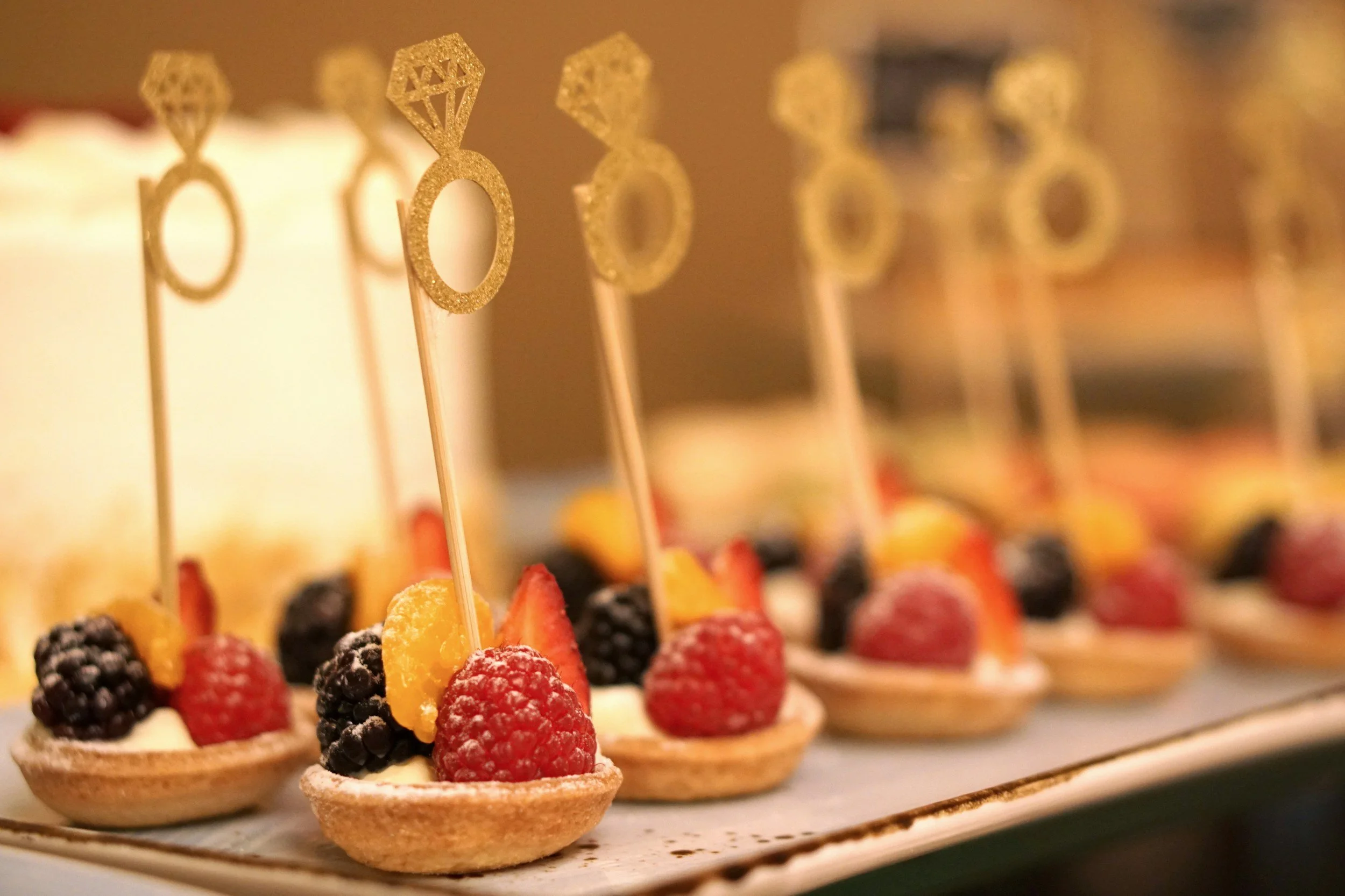 Mini fruit tarts topped with blackberries, raspberries, strawberries, and slices of yellow fruit, each with a glittery gold ring-shaped pick with a diamond on top.