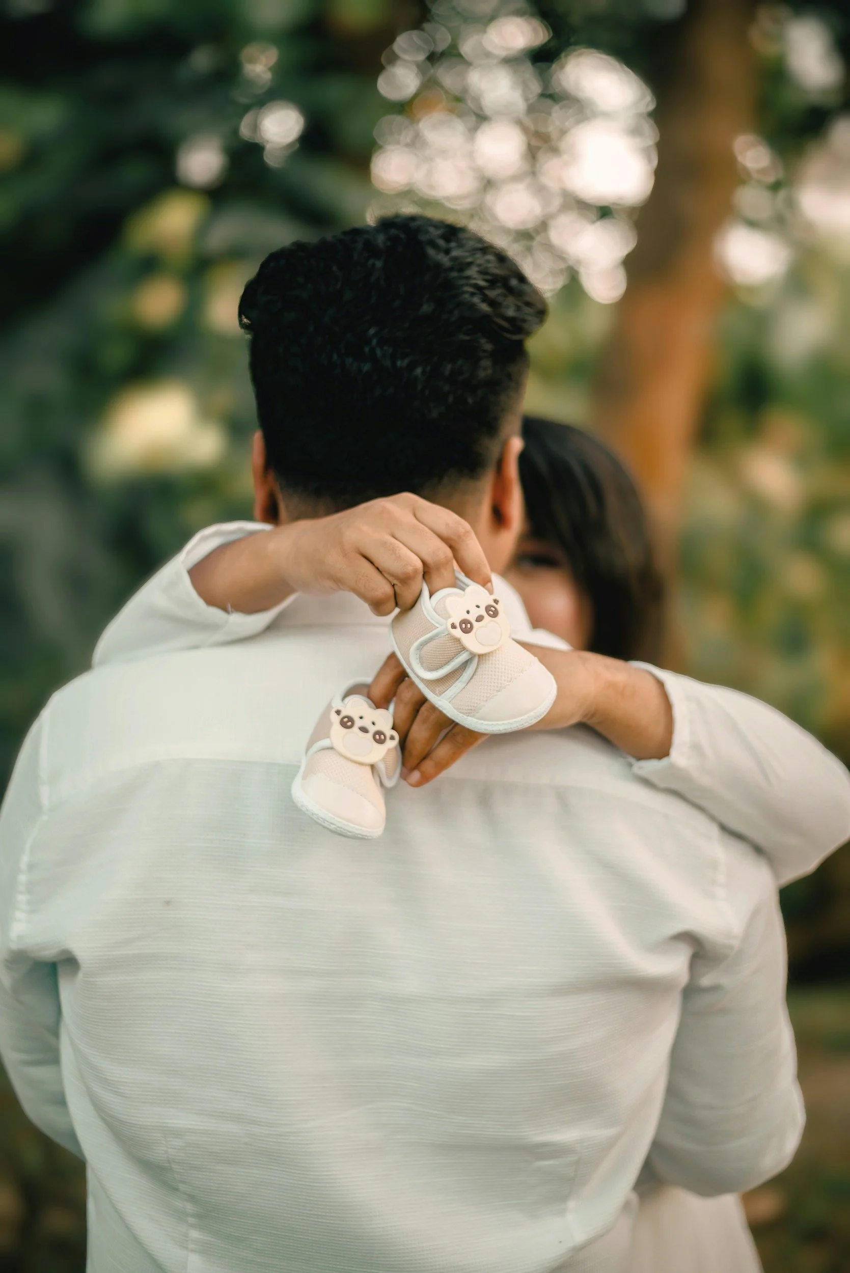 A person holds a small baby shoe decorated with a cute bear face over their shoulder, with another person partially visible in the background, outdoors with blurred greenery.