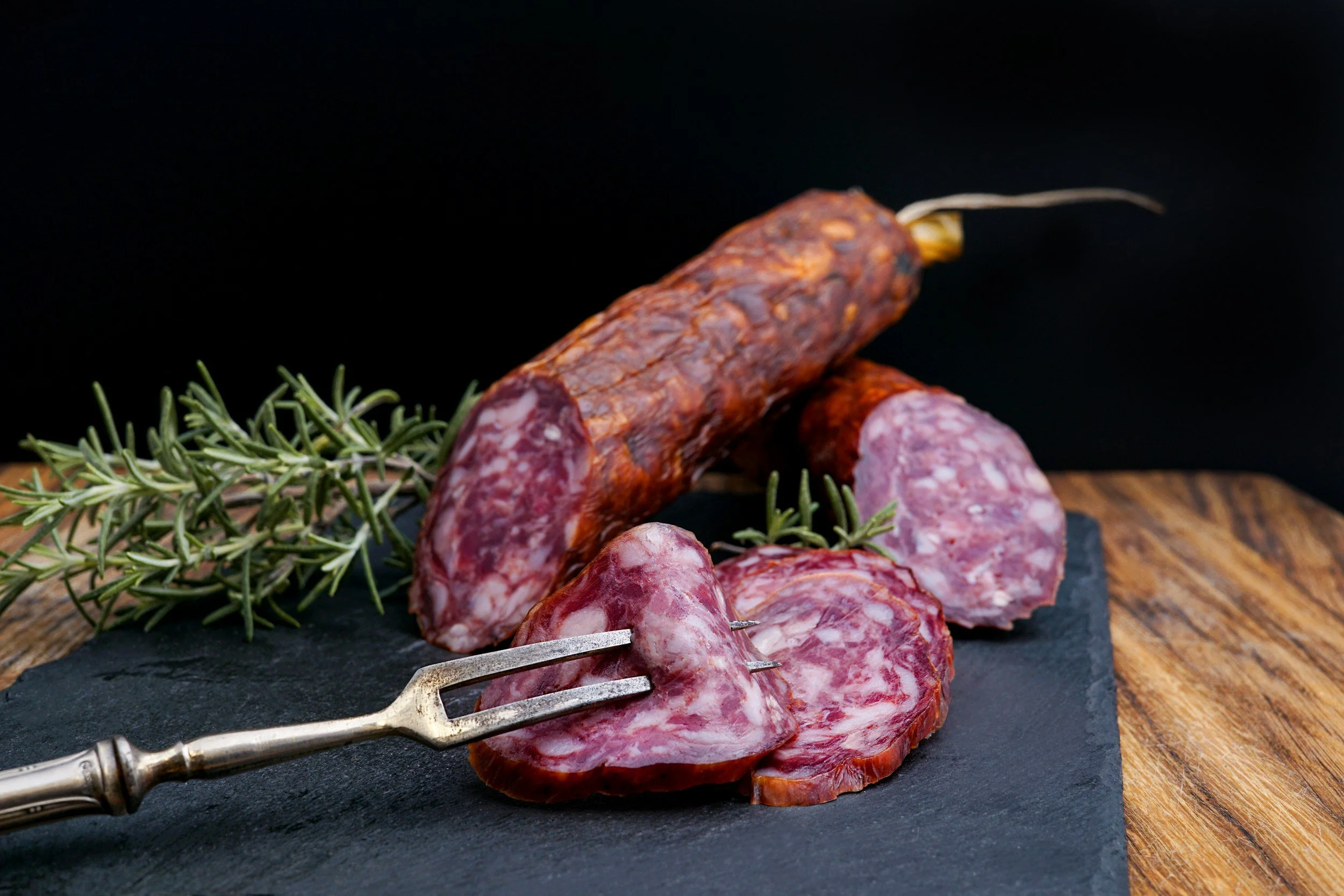 Sliced cured sausage on slate board with sprigs of rosemary, dark background