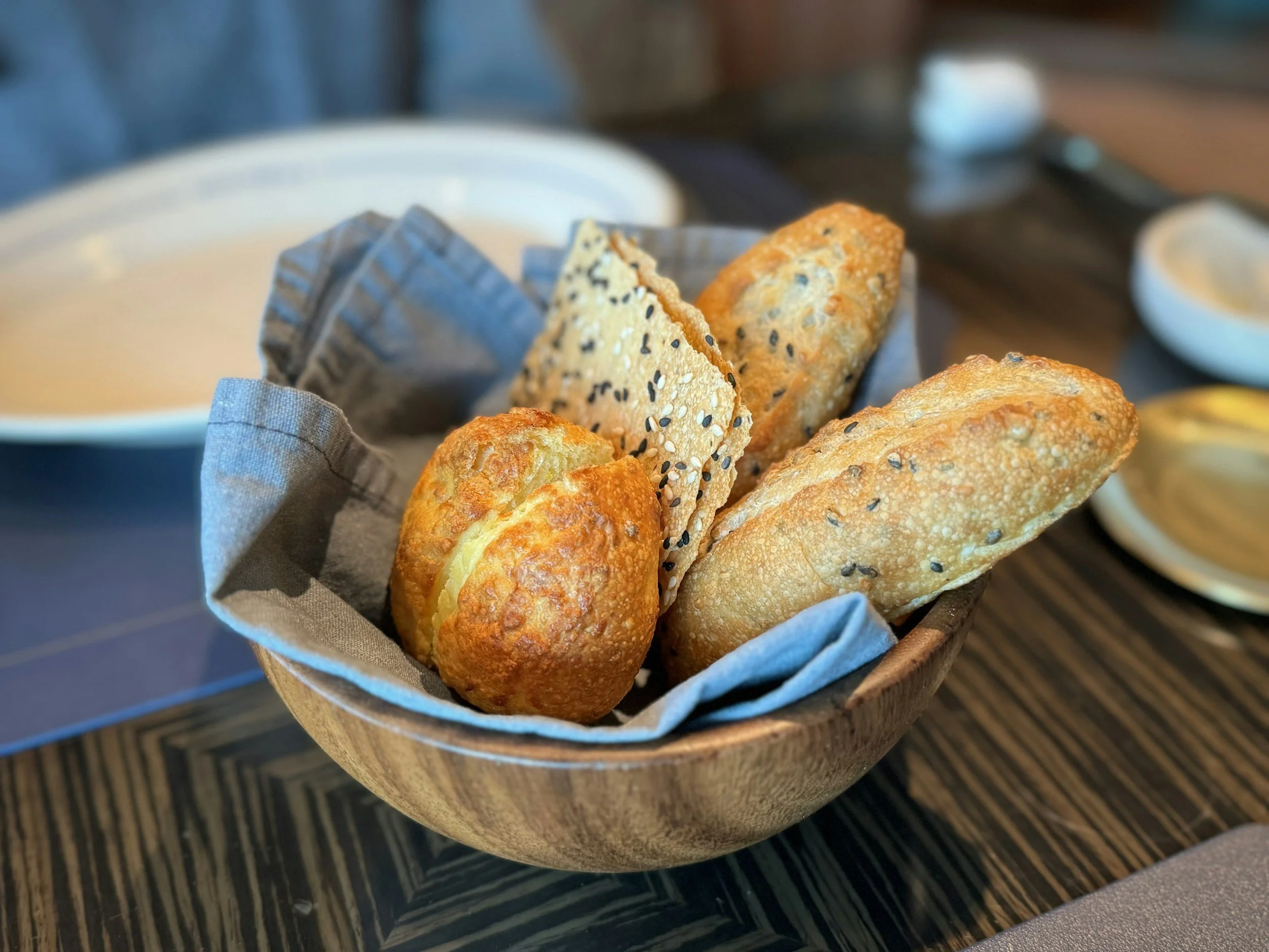 A wicker basket bowl with assorted breadsticks and crackers, lined with parchment paper, on a wooden table with small dishes of dipping oils in the background.