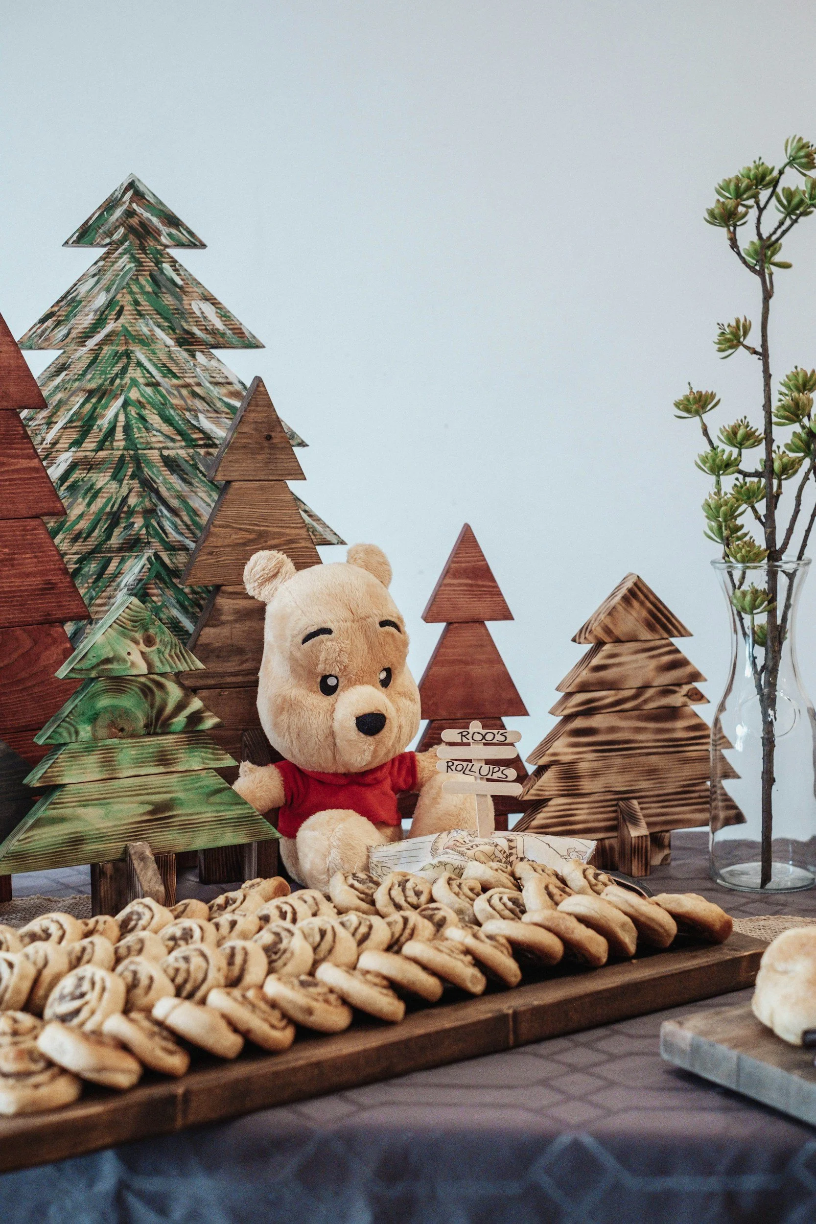 A Winnie the Pooh plush toy sitting in front of a wooden forest scene with trees and a sign that reads 'Roo's Roll-Ups'. There is a tray of swirled cookies in the foreground and a tall glass vase with a succulent plant on the right.