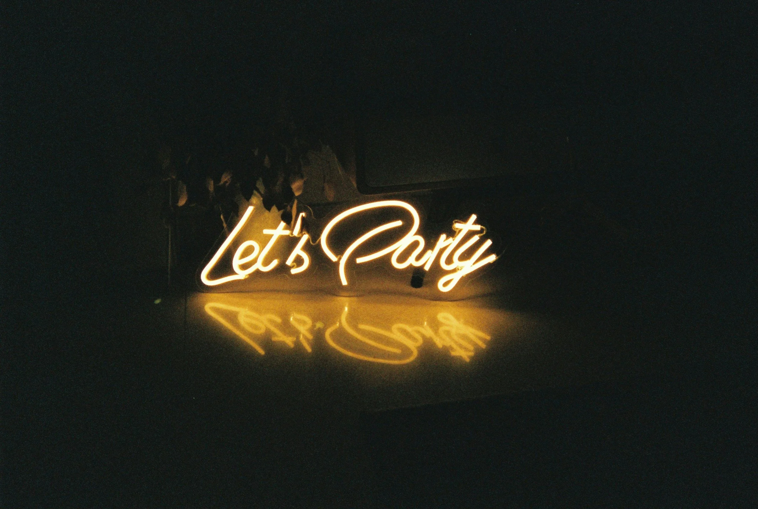 Neon sign with the words "Let's Party" glowing in yellow, reflecting on a dark surface.