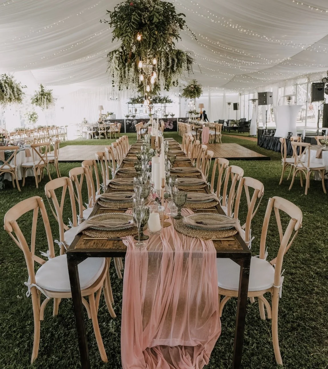 Elegant wedding reception setup inside a large white tent with string lights, long wooden tables with pink table runners, candle centerpieces, and wooden chairs with white cushions.