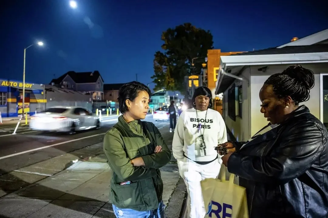 Three women standing on a city sidewalk at night, with moving cars and buildings in the background. One woman is wearing a green jacket, another is in a white hoodie that says "BISOUS PARIS," and the third woman is in a black leather jacket, looking at her phone.