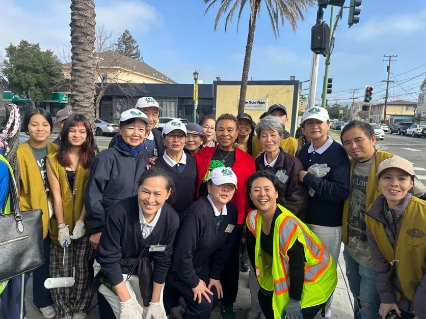 Group of diverse volunteers gathered outdoors at an intersection, some wearing yellow vests and caps, with palm trees and traffic lights in the background.