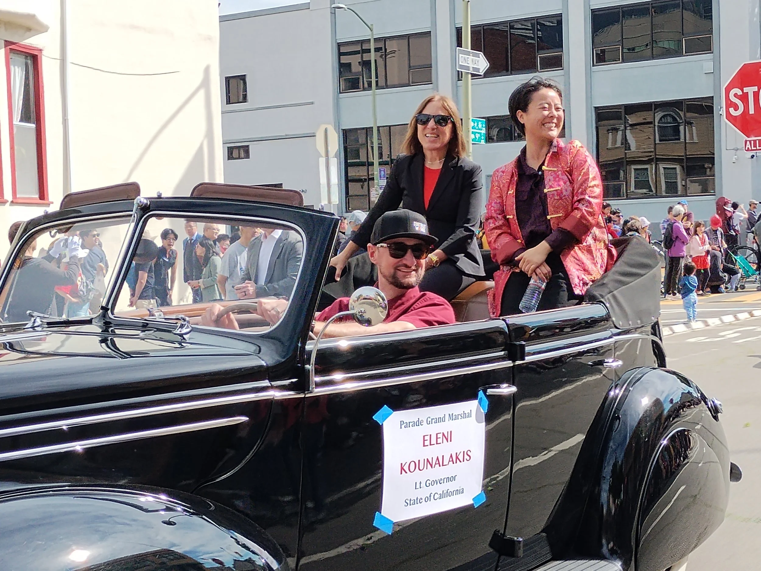Two women riding in a classic black convertible car during a parade. The woman on the left has shoulder-length hair, wears sunglasses, a black blazer, and a red top, and is smiling. The woman on the right has short hair, wears a patterned pink and orange jacket, and is smiling. Behind the car, crowd of parade spectators can be seen, along with a sign that reads 'Parade Grand Marshal' and 'Eleni Kounalakis, Lt Governor of California' on the side of the car. The background shows city buildings, a street sign, and a stop sign.