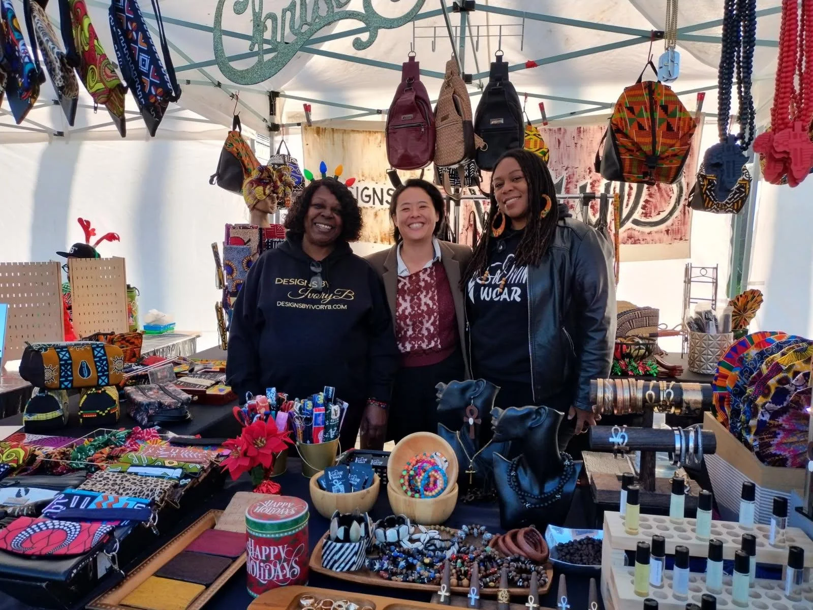 Three women standing behind a stall at a craft fair, displaying a variety of colorful handmade jewelry, accessories, and bags, with African-inspired patterns. The background features more bags hanging from the tent and a sign that reads 'Designs by Ivory B'.