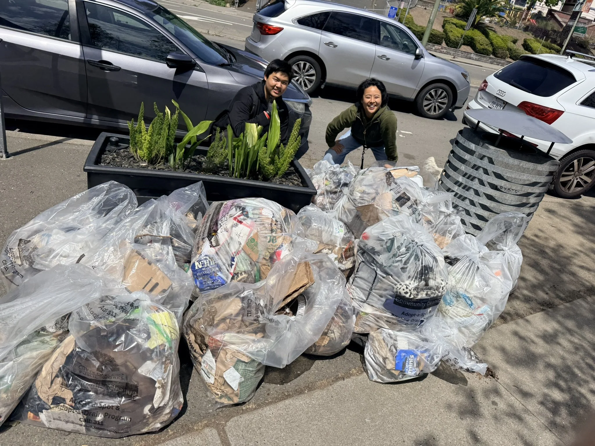 Two people crouching next to a large pile of trash bags filled with recyclables on a street sidewalk with parked cars and some greenery in the background.