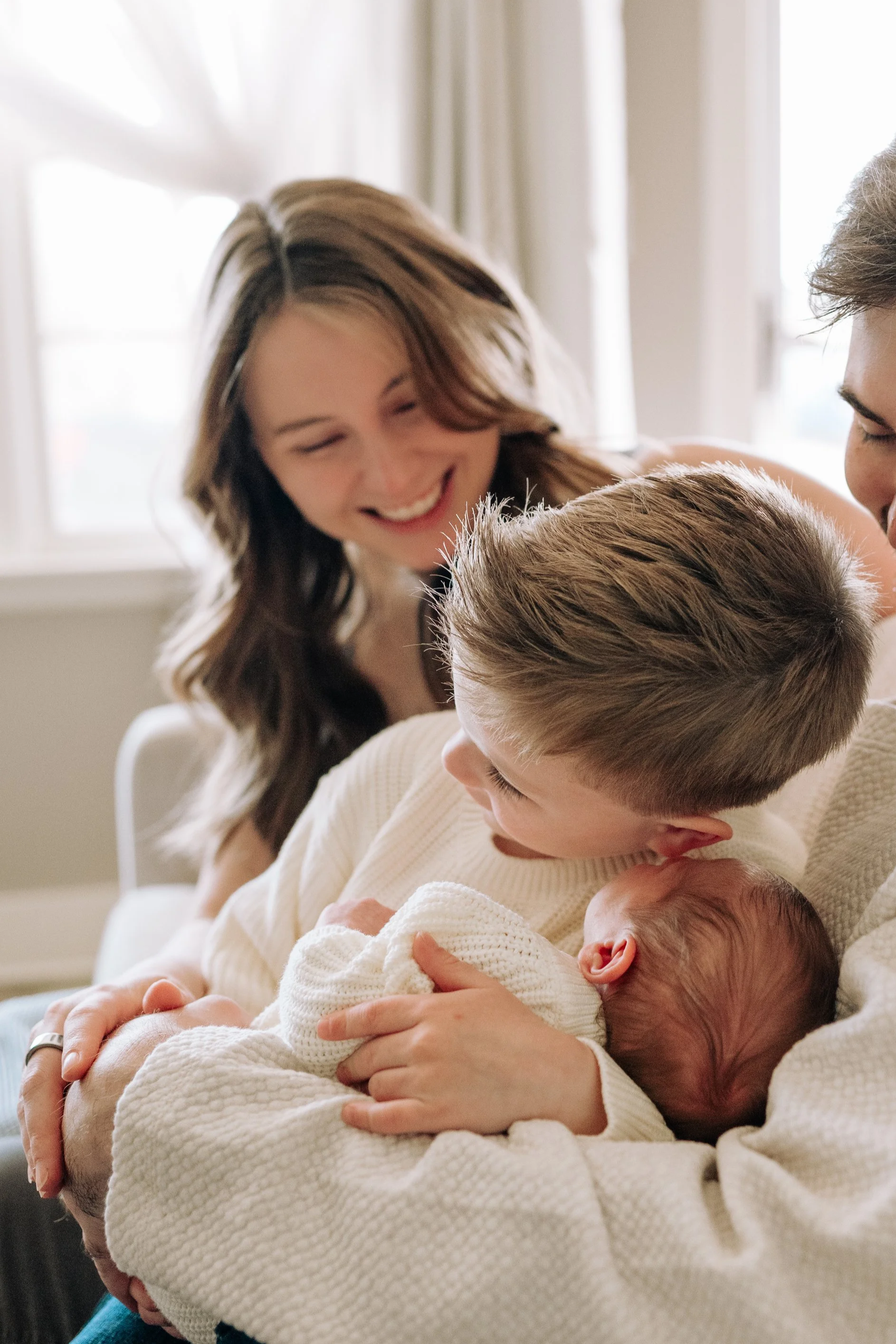 A family with a woman, a young boy, and a man holding a newborn baby, smiling and gazing at the baby in a warmly lit room.