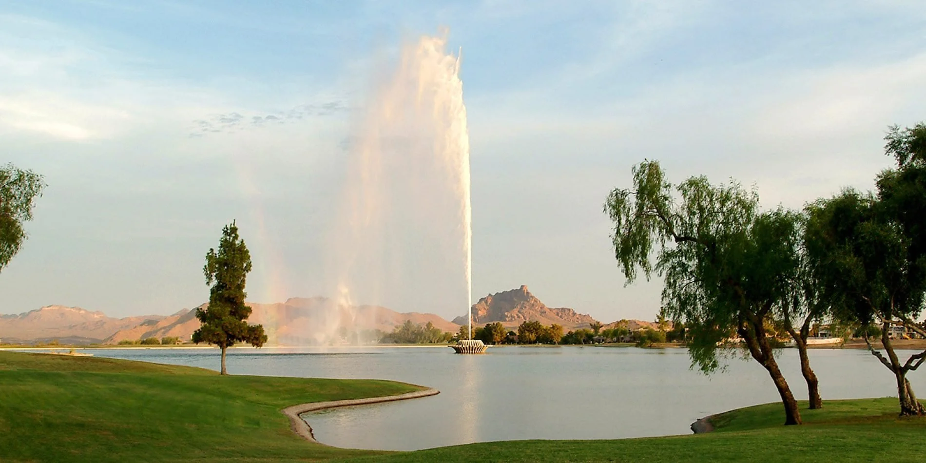 A large fountain in the center of the image shoots a jet of water high in the air. The fountain is in a small lake. The foreground consists of manicured grass, while the background shows rugged mountain peaks.